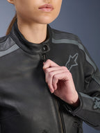 Kurtka skórzana Alpinestars Stella Dyno, zbliżenie na przedni zamek i kołnierzyk regulowany przez modelkę, czarna z akcentami w kolorze Tar Gray i ciemnoszarym, z haftowanym logo Alpinestars na piersi, tekstura wysokiej jakości skóry bydlęcej, miejska odzież motocyklowa dla kobiet
