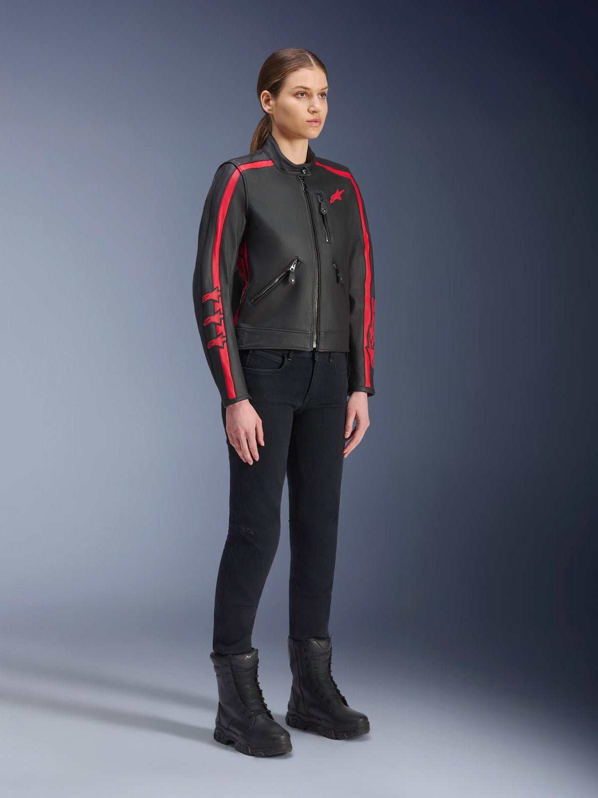 Modelka w skórzanej kurtce Alpinestars Stella Dyno w kolorze Black Haute Red, zestawionej z czarnymi spodniami i butami, skórzana kurtka motocyklowa w stylu heritage z czerwonymi pasami na rękawach i czerwonym logo, damska odzież motocyklowa do jazdy miejskiej i szosowej