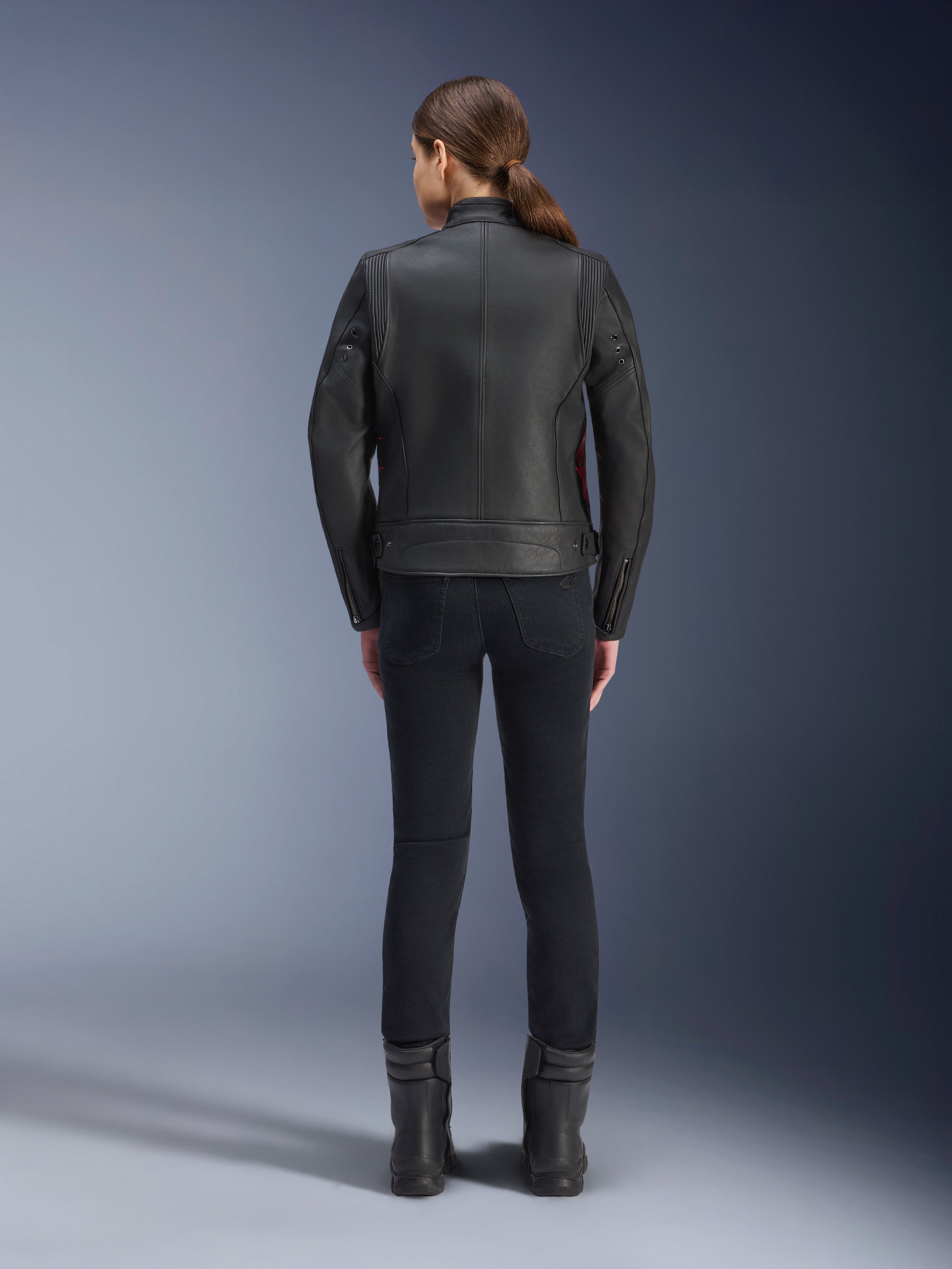 Widok z tyłu kobiety ubranej w skórzaną kurtkę Alpinestars Stella Dyno, czarna z akcentami Haute Red, w zestawieniu z dopasowanymi czarnymi spodniami i czarnymi butami, nowoczesny design skórzanej kurtki motocyklowej typu heritage.