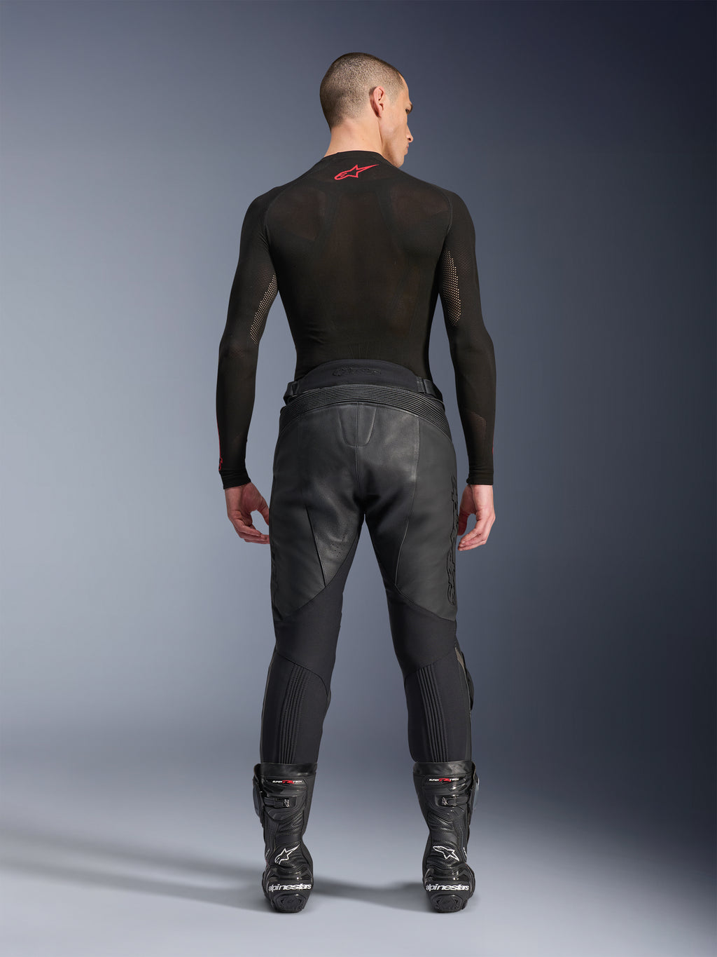 Alpinestars Missile V3 Leather Pants, czarne, widok z tyłu na modelu ubranym w czarną koszulkę kompresyjną z długim rękawem i buty wyścigowe, konstrukcja z wysokiej jakości skóry z podwyższonym stanem i strefami elastycznymi, profesjonalna odzież motocyklowa wyścigowa