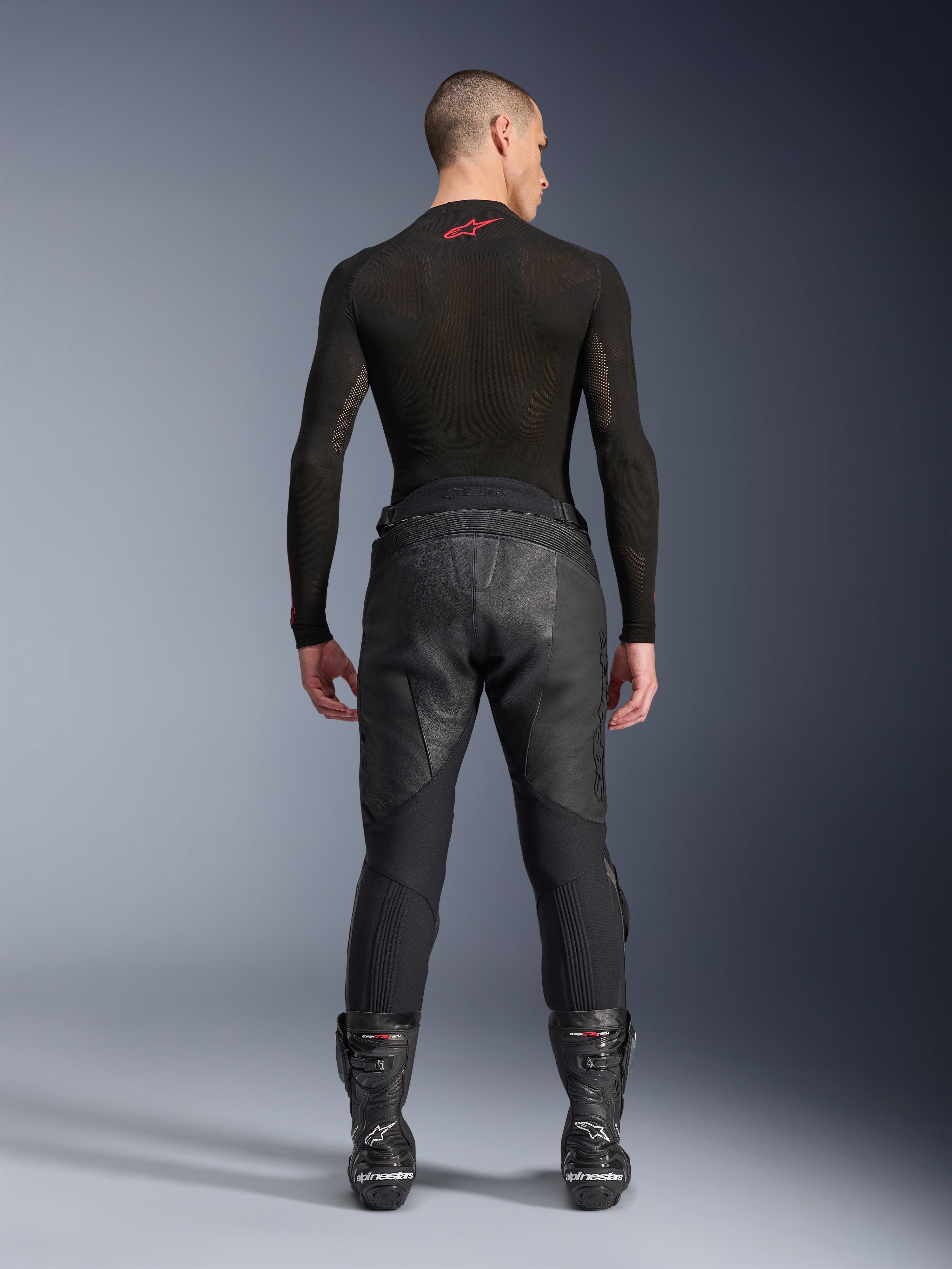 Alpinestars Missile V3 Leather Pants, czarne, widok z tyłu na modelu ubranym w czarną koszulkę kompresyjną z długim rękawem i buty wyścigowe, konstrukcja z wysokiej jakości skóry z podwyższonym stanem i strefami elastycznymi, profesjonalna odzież motocyklowa wyścigowa