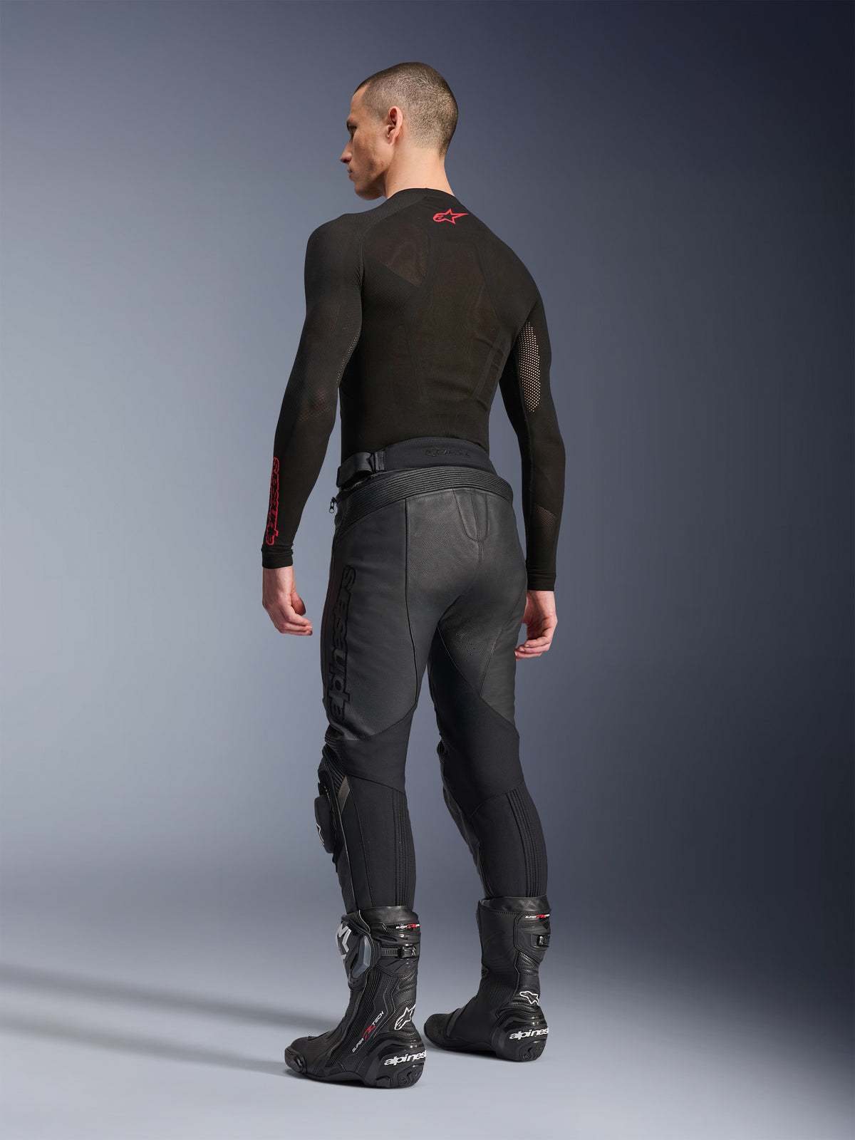 Skórzane spodnie Alpinestars Missile V3, czarne, widok z tyłu na modelu, noszone z czarną warstwą bazową z długim rękawem i czarnymi motocyklowymi butami wyścigowymi, wyposażone w harmonijkowe skórzane panele elastyczne i wstępnie zakrzywioną konstrukcję dla ergonomicznego dopasowania podczas jazdy.