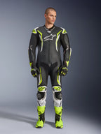 Alpinestars GP Tech V5 One Piece Leather Suit, profesjonalny motocyklowy kombinezon wyścigowy w kolorze czarnym i neonowo-żółtym fluo z białymi akcentami, noszony przez modela z dopasowanymi rękawicami i butami wyścigowymi, o aerodynamicznym designie i z ochronnymi sliderami.