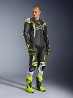 Alpinestars GP Tech V5 One Piece Leather Suit, czarno-biało-żółty fluo, prezentowany przez modela z dopasowanymi rękawicami i butami wyścigowymi, profesjonalny motocyklowy ubiór wyścigowy o właściwościach aerodynamicznych z ochronnymi ochraniaczami.