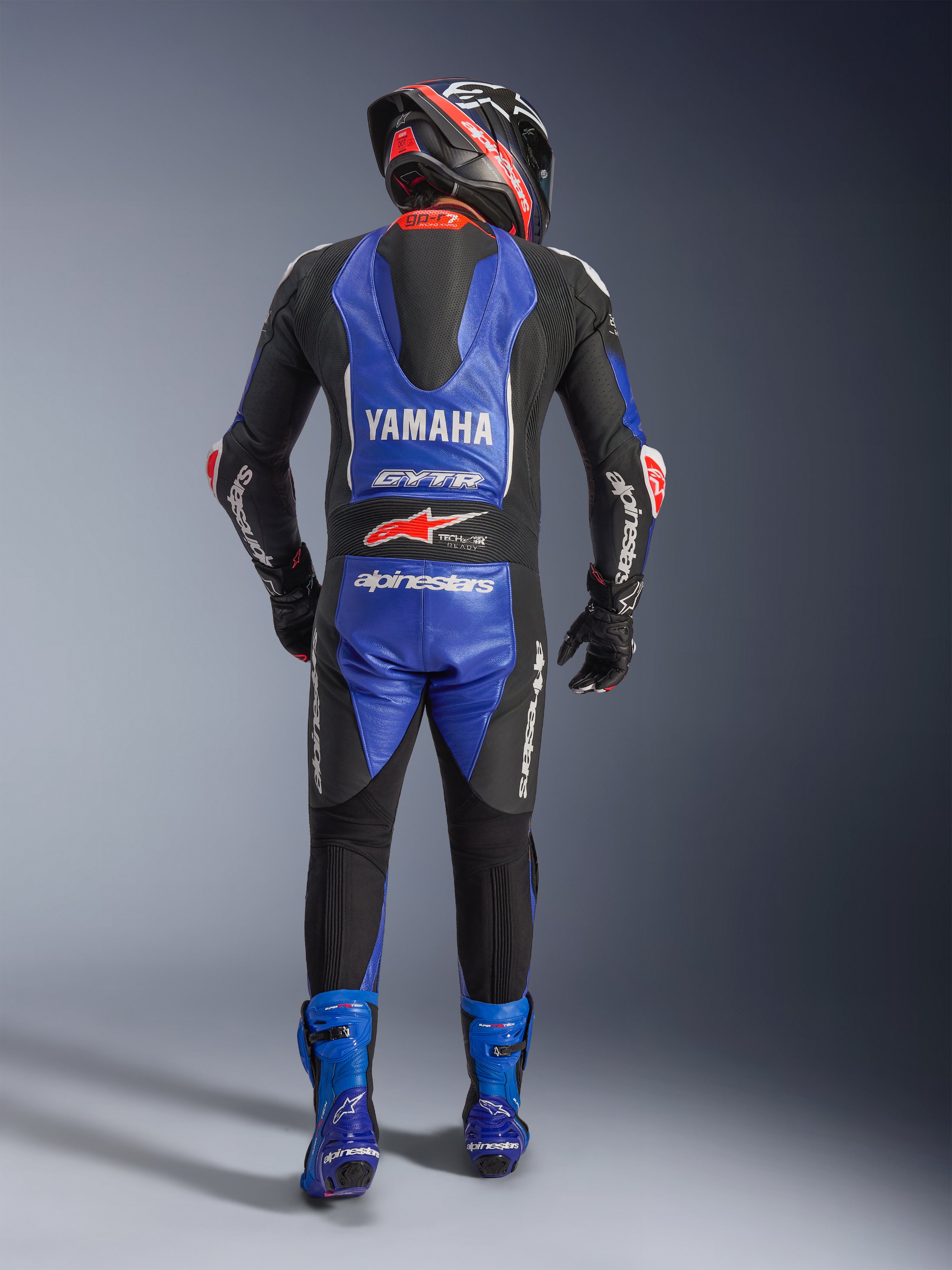 Alpinestars Yamaha GP-R7 One Piece Leather Suit, widok z tyłu motocyklisty w pełnym rynsztunku wyścigowym, metaliczny niebieski i czarny z białymi i czerwonymi akcentami, z logo Yamaha i GYTR na garbie aerodynamicznym, noszony z pasującymi niebieskimi butami i kaskiem wyścigowym.
