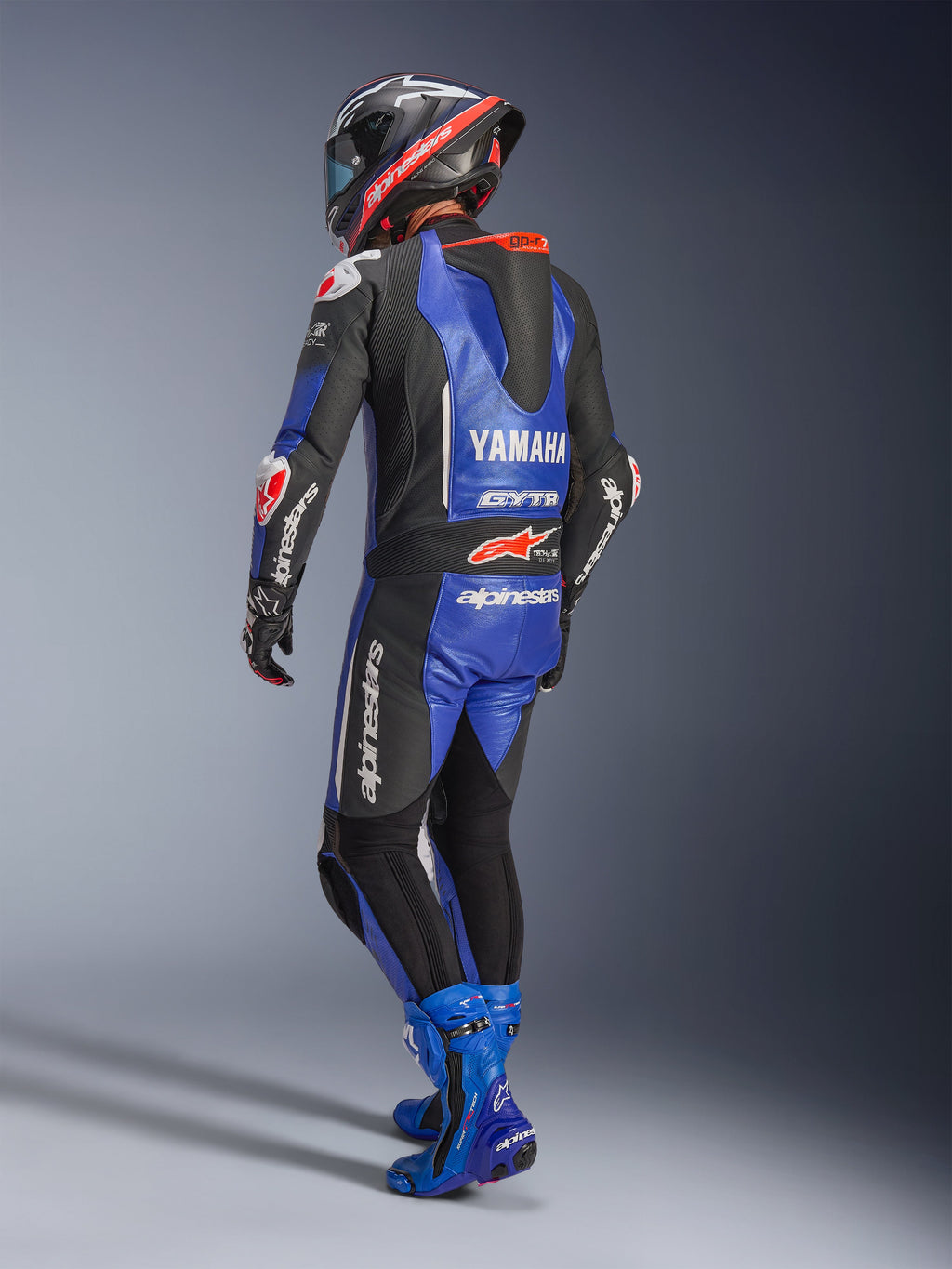 Jednoczęściowy kombinezon skórzany Alpinestars Yamaha GP-R7, metaliczny niebieski, czarny i biały, widok z tyłu pokazujący aerodynamiczny garb wyścigowy i branding Yamaha, noszony z dopasowanymi niebieskimi butami wyścigowymi i kaskiem, profesjonalny wyczynowy kombinezon skórzany do wyścigów torowych