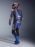 Jednoczęściowy kombinezon skórzany Alpinestars Yamaha GP-R7, metaliczny niebieski, czarny i biały, widok z tyłu pokazujący aerodynamiczny garb wyścigowy i branding Yamaha, noszony z dopasowanymi niebieskimi butami wyścigowymi i kaskiem, profesjonalny wyczynowy kombinezon skórzany do wyścigów torowych