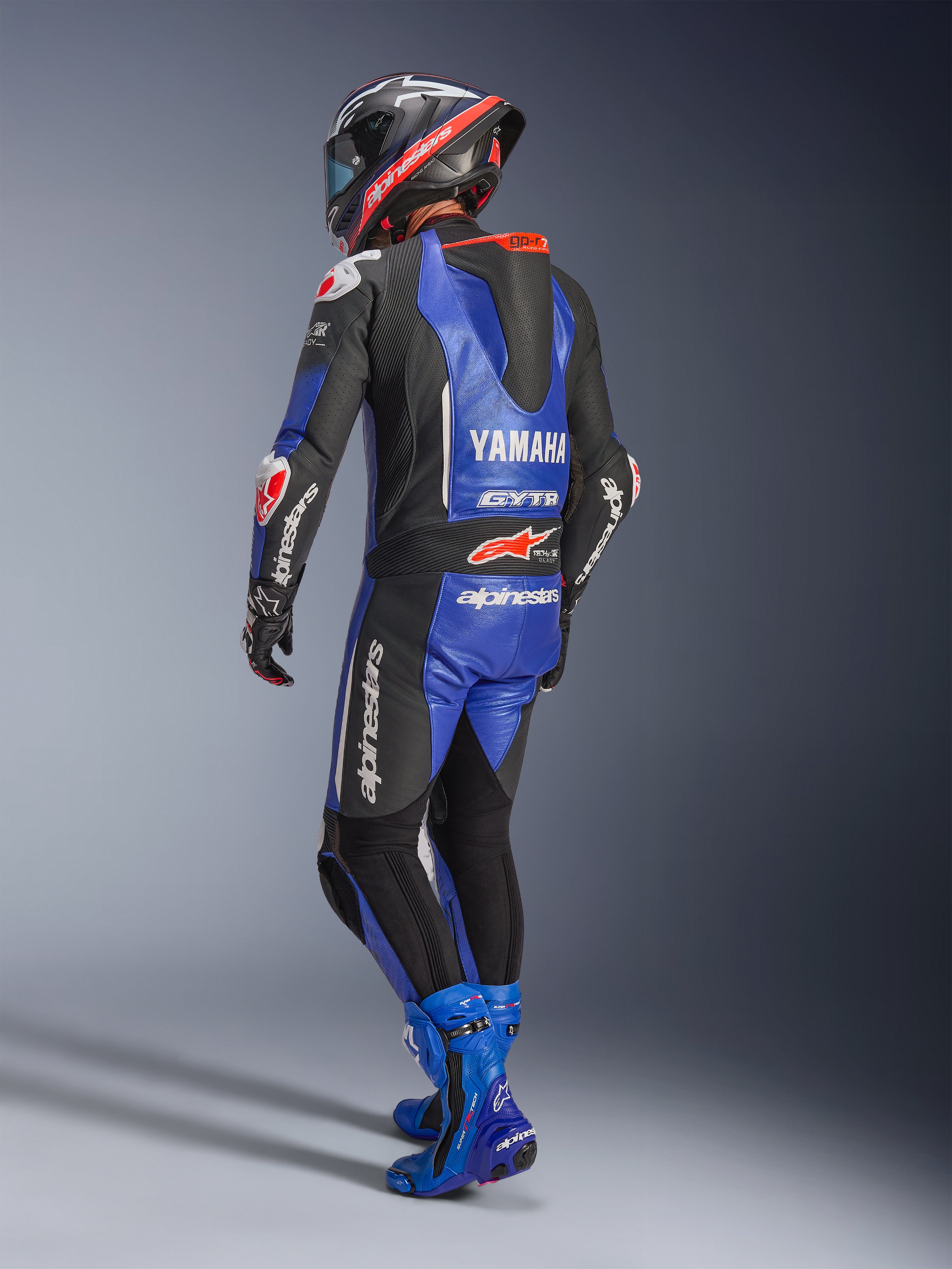 Jednoczęściowy kombinezon skórzany Alpinestars Yamaha GP-R7, metaliczny niebieski, czarny i biały, widok z tyłu pokazujący aerodynamiczny garb wyścigowy i branding Yamaha, noszony z dopasowanymi niebieskimi butami wyścigowymi i kaskiem, profesjonalny wyczynowy kombinezon skórzany do wyścigów torowych