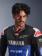 Portret z bliska mężczyzny w jednoczęściowym kombinezonie skórzanym Alpinestars Yamaha GP-R7, metaliczny niebieski i czarny z białym brandingiem YAMAHA i GYTR, perforowana skóra, białe slidery na ramionach z czerwonym logo gwiazdy, Tech-Air Ready, profesjonalna odzież motocyklowa