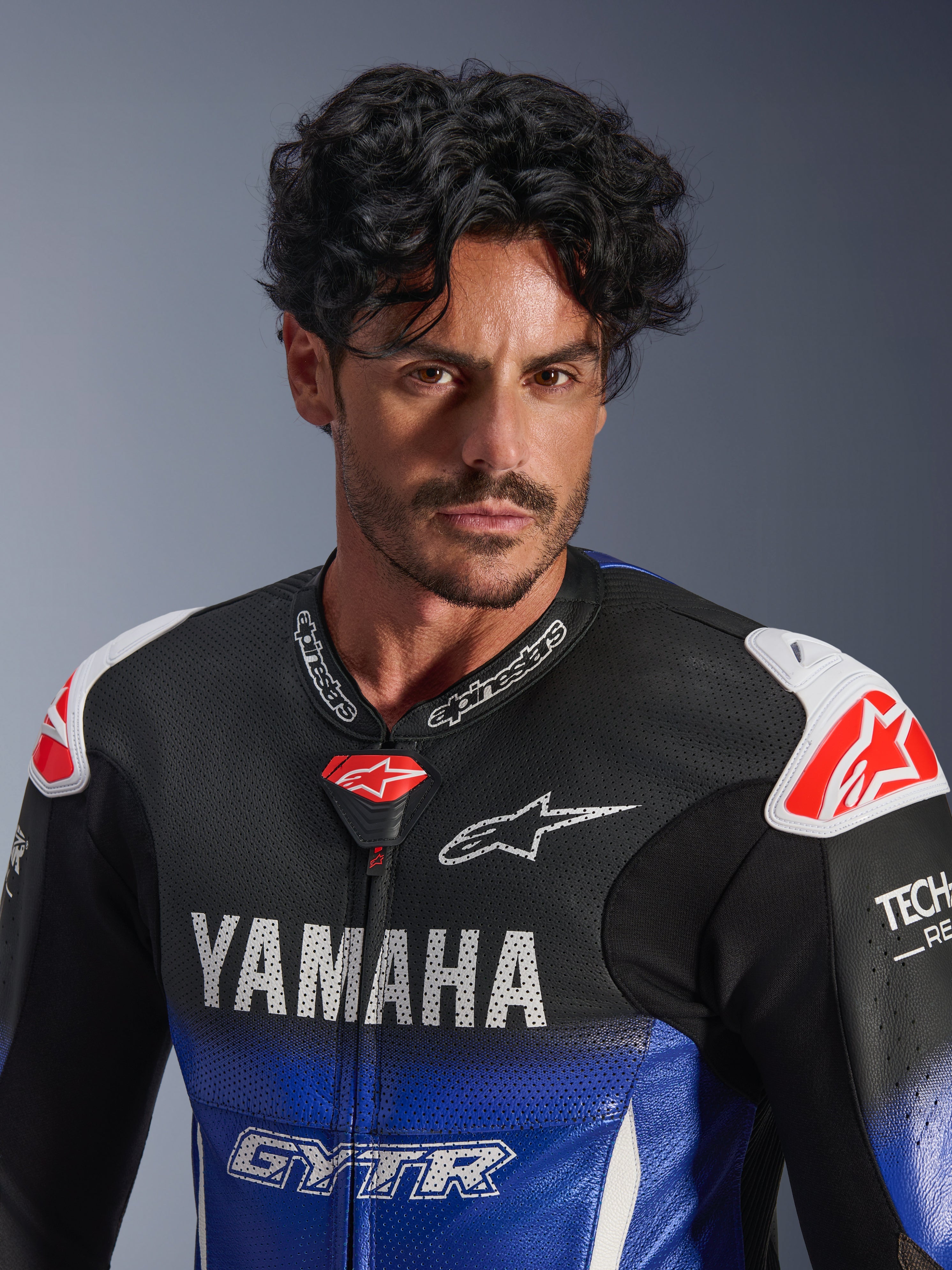 Portret z bliska mężczyzny w jednoczęściowym kombinezonie skórzanym Alpinestars Yamaha GP-R7, metaliczny niebieski i czarny z białym brandingiem YAMAHA i GYTR, perforowana skóra, białe slidery na ramionach z czerwonym logo gwiazdy, Tech-Air Ready, profesjonalna odzież motocyklowa