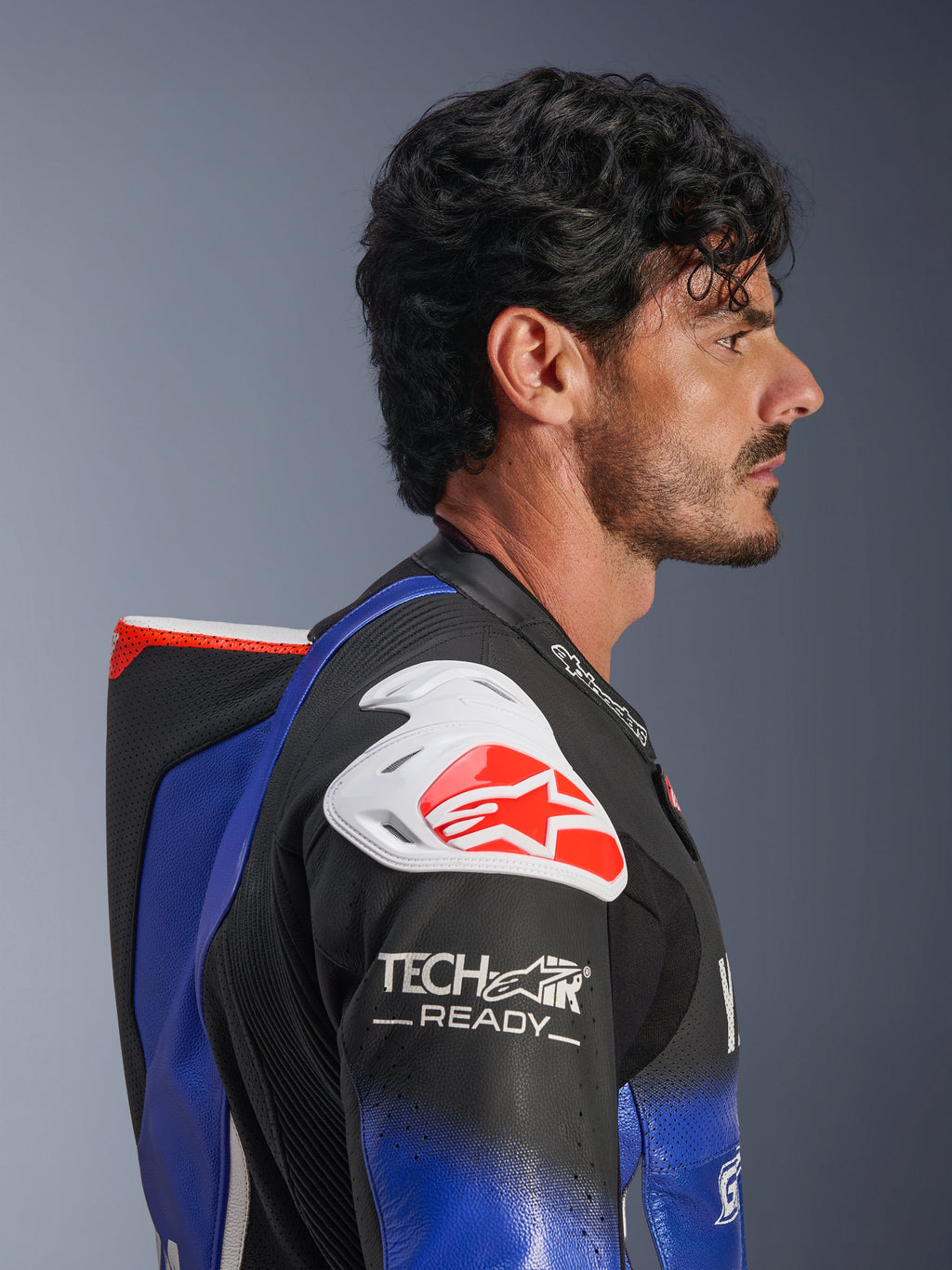 Jednoczęściowy kombinezon skórzany Alpinestars Yamaha GP-R7, metaliczny niebieski i czarny z białymi i czerwonymi akcentami, profil boczny podkreślający aerodynamiczny garb i biały ochraniacz ramienia z czerwonym logo Alpinestars, oznaczenie Tech-Air Ready na rękawie, najwyższej jakości perforowana skóra wyścigowa.