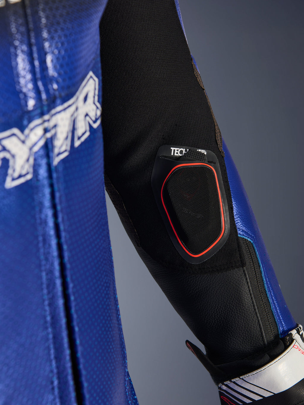 Zbliżenie na przedramię jednoczęściowego kombinezonu skórzanego Yamaha GP-R7, ze zintegrowaną kieszenią na wyświetlacz Bluetooth Tech-Air 10 z czerwoną oblamówką, metaliczną niebieską perforowaną skórą i czarnymi wstawkami z elastycznej tkaniny dla wyścigowych osiągów.