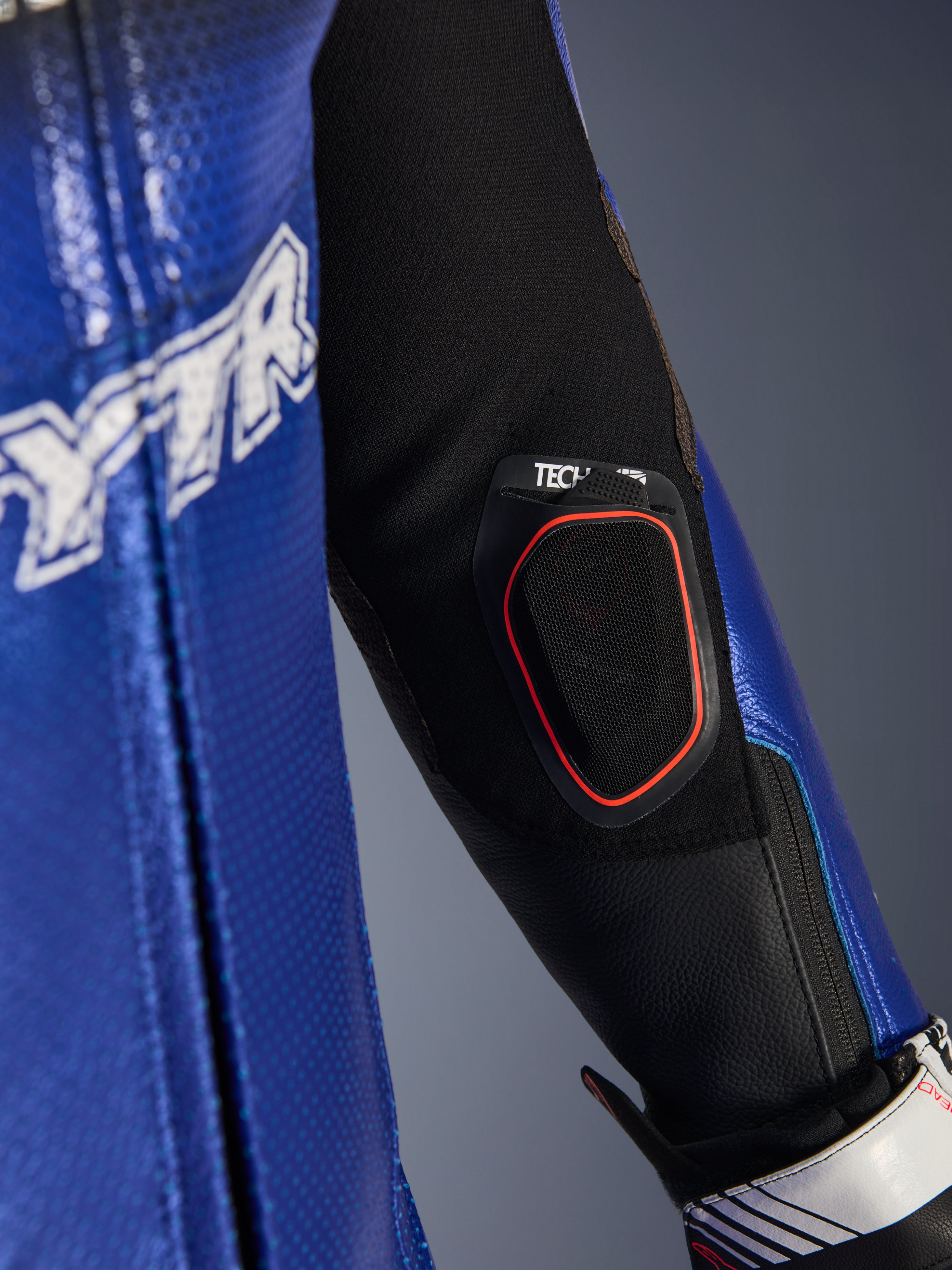 Zbliżenie na przedramię jednoczęściowego kombinezonu skórzanego Yamaha GP-R7, ze zintegrowaną kieszenią na wyświetlacz Bluetooth Tech-Air 10 z czerwoną oblamówką, metaliczną niebieską perforowaną skórą i czarnymi wstawkami z elastycznej tkaniny dla wyścigowych osiągów.