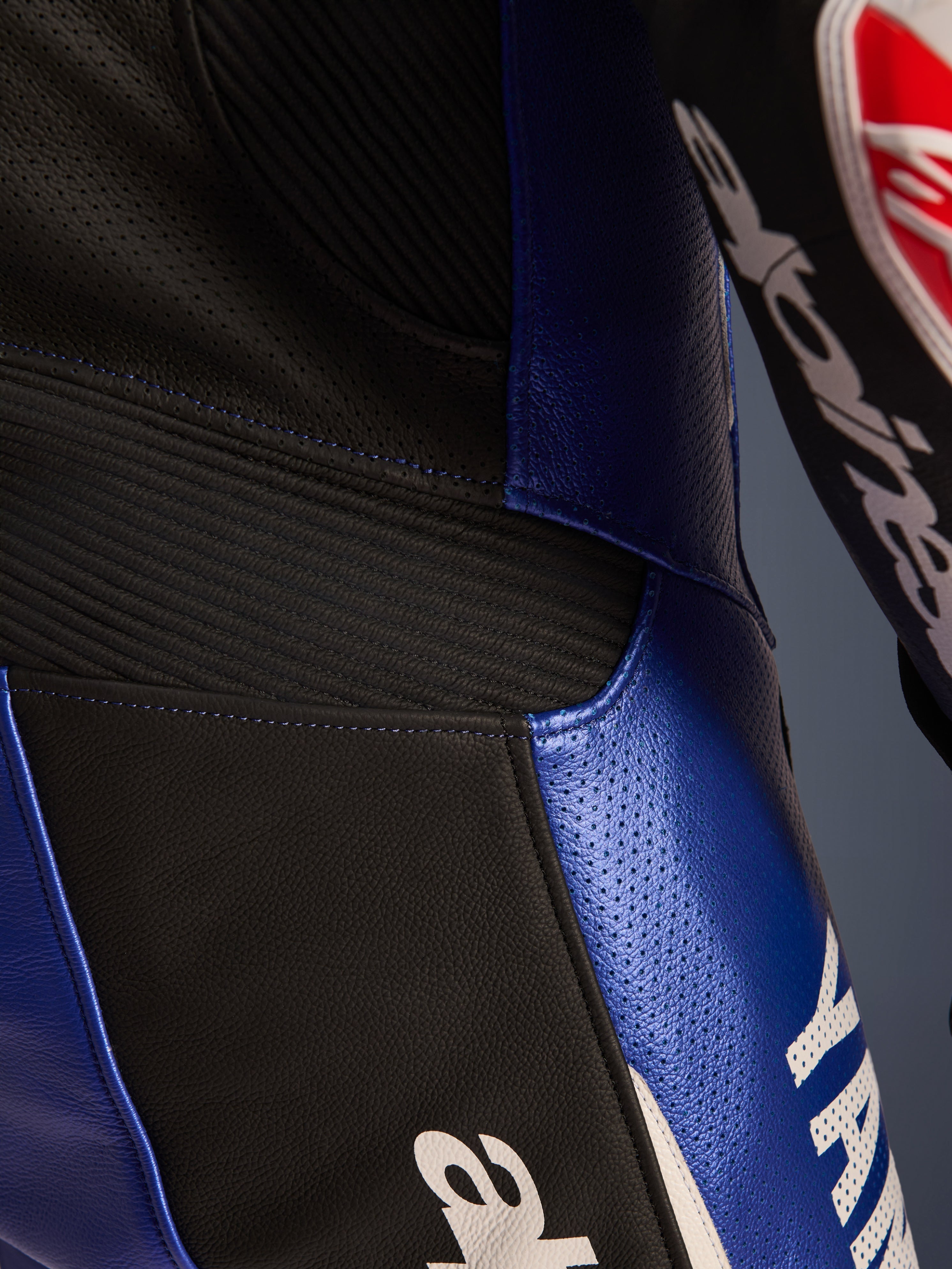 Zbliżenie na detale jednoczęściowego kombinezonu skórzanego Alpinestars Yamaha GP-R7, niebieski metalik i czarny, z perforowanymi panelami skórzanymi zapewniającymi wentylację, czarnymi elastycznymi wstawkami akordeonowymi i wyraźnym białym logo Yamaha, podkreślające konstrukcję z wyścigowej skóry bydlęcej.