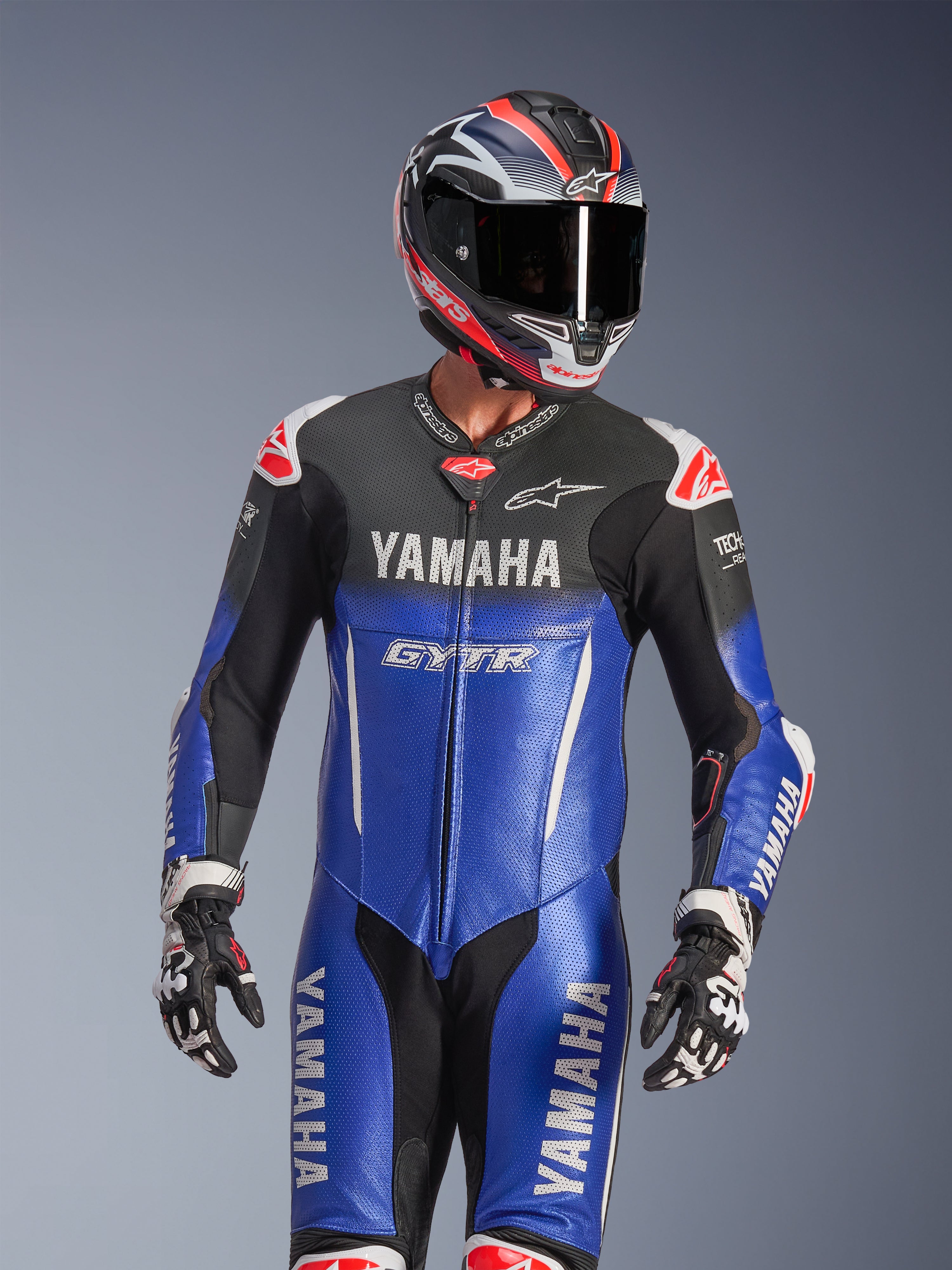 Kombinezon skórzany Yamaha GP-R7 1PC, metaliczny niebieski i czarny z białymi i czerwonymi akcentami, noszony z dopasowanym kaskiem i rękawicami wyścigowymi, branding YAMAHA i GYTR, perforowane panele skórzane oraz logo Alpinestars, profesjonalna odzież motocyklowa.