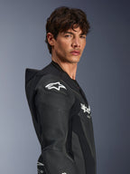 Męski model w jednoczęściowym skórzanym kombinezonie Alpinestars GP Force V2 w kolorze czarnym, widok z profilu eksponujący białe logo gwiazdy Alpinestars na protektorze ramienia DFS Lite, perforowane panele skórzane oraz aerodynamiczny garb prędkości.