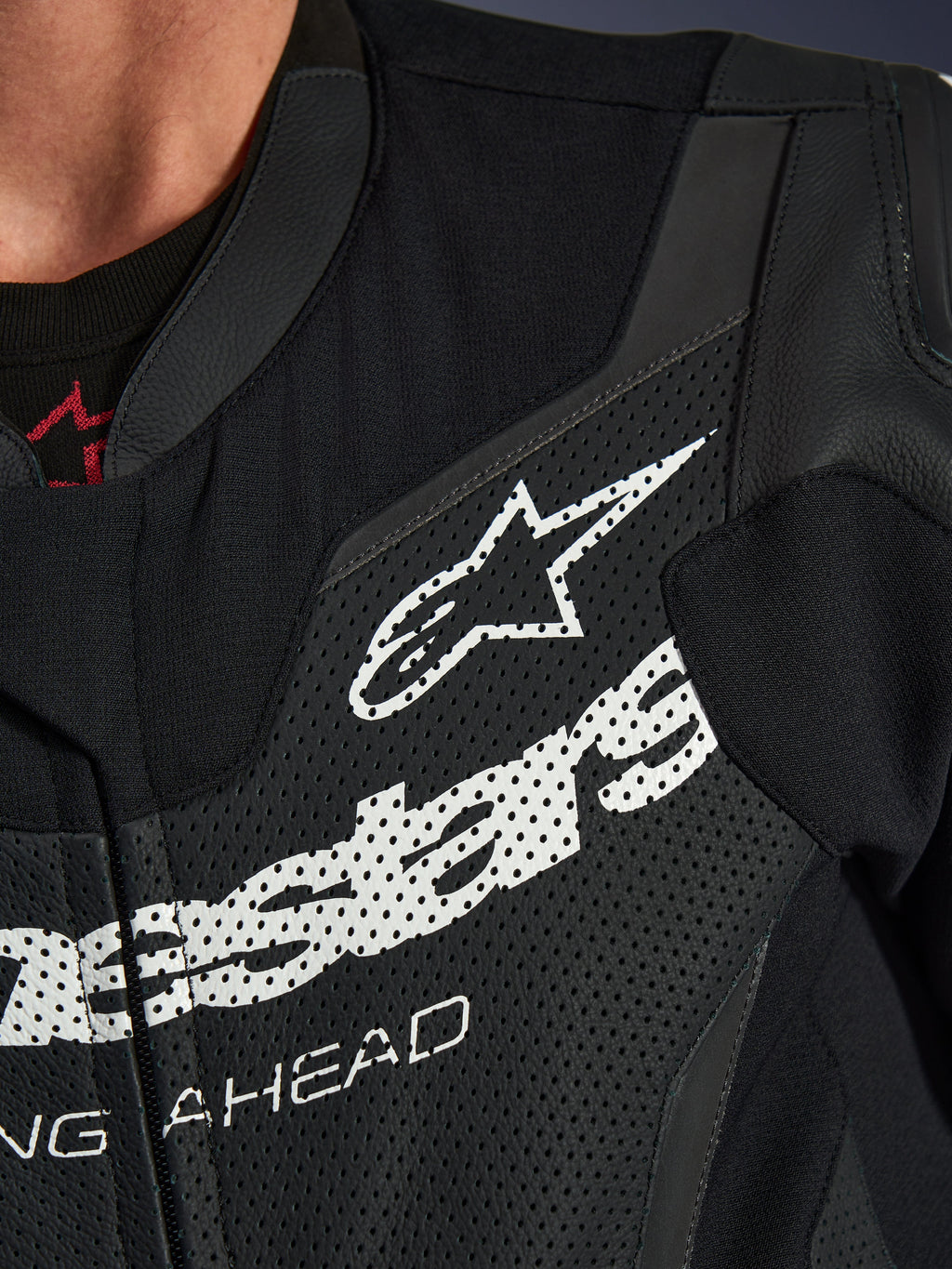 Jednoczęściowy kombinezon skórzany Alpinestars GP Force V2, zbliżenie na klatkę piersiową, czarna perforowana skóra z dużym białym logo Alpinestars, techniczna sportowa odzież motocyklowa, szczegółowa tekstura i branding