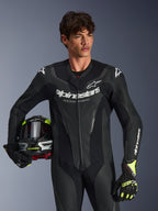 Kombinezon skórzany Alpinestars GP Force V2, jednoczęściowy czarny motocyklowy kombinezon wyścigowy z perforowanymi panelami skórzanymi i białym logo, noszony przez modela trzymającego kask i pasujące rękawice, oświetlenie studyjne
