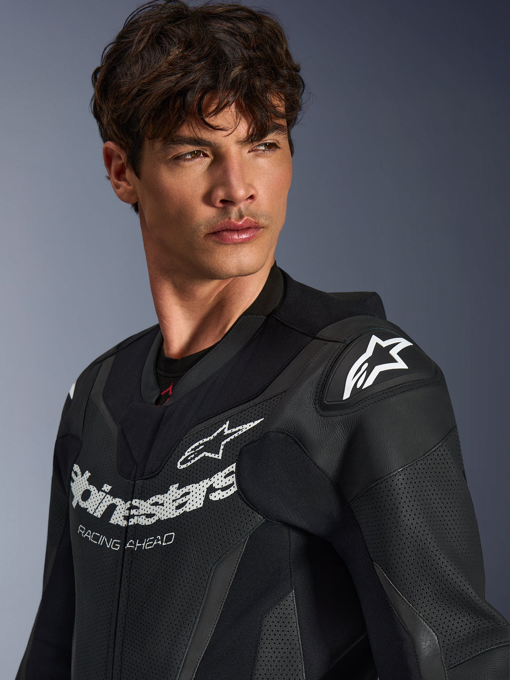 Zbliżenie na mężczyznę ubranego w czarny skórzany kombinezon Alpinestars GP Force V2, z perforowaną konstrukcją skórzaną, logo Alpinestars z napisem „RACING AHEAD” na klatce piersiowej oraz białym logo gwiazdy na ślizgaczach ramion.