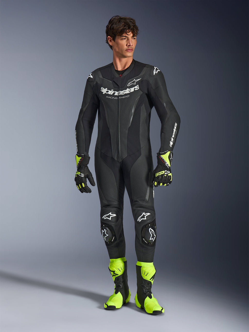 Alpinestars GP Force V2 Leather Suit, jednoczęściowy czarny motocyklowy kombinezon wyścigowy, noszony z neonowożółtymi butami i rękawicami, z białymi logo i perforowaną skórą, dopasowany krój na dni torowe