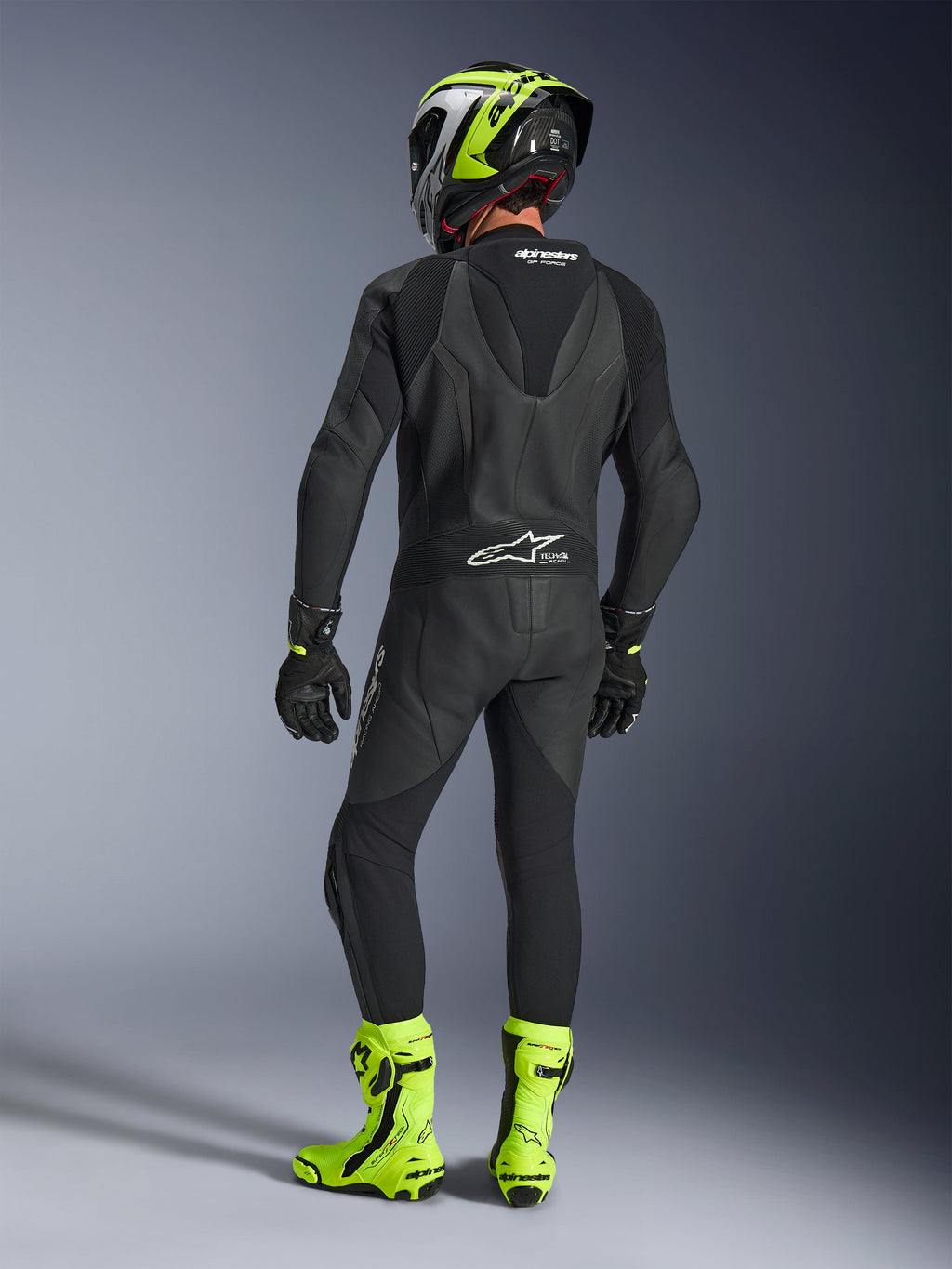 Widok z tyłu motocyklisty w jednoczęściowym skórzanym kombinezonie Alpinestars GP Force V2 w kolorze czarnym, prezentujący aerodynamiczny garb i logo Tech-Air Ready, w zestawie z czarno-neonowożółtym kaskiem oraz jaskrawożółtymi butami wyścigowymi.