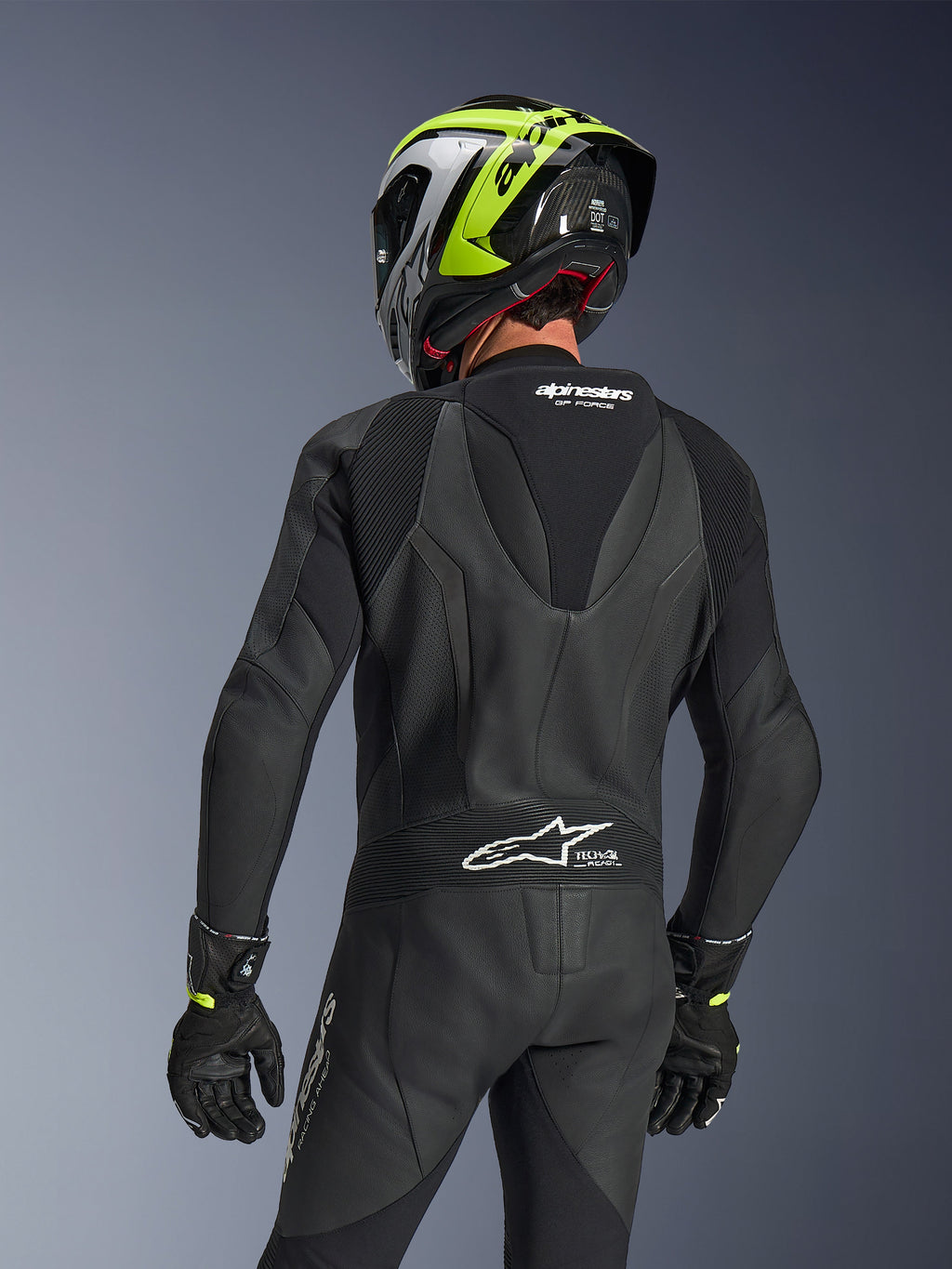 Alpinestars GP Force V2 Leather Suit 1-Piece, czarny, widok z tyłu przedstawiający garb aerodynamiczny i perforowaną konstrukcję skórzaną, zestawiony z czarno-neonowożółtym kaskiem wyścigowym i czarnymi rękawicami, harmonijkowe panele elastyczne i białe logo Tech-Air Ready