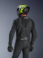 Alpinestars GP Force V2 Leather Suit 1-Piece, czarny, widok z tyłu przedstawiający garb aerodynamiczny i perforowaną konstrukcję skórzaną, zestawiony z czarno-neonowożółtym kaskiem wyścigowym i czarnymi rękawicami, harmonijkowe panele elastyczne i białe logo Tech-Air Ready