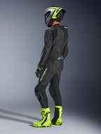 Alpinestars GP Force V2 Leather Suit 1-Piece, czarny jednoczęściowy kombinezon skórzany, widok z tyłu i z boku, białe logo na udzie i dolnej części pleców, aerodynamiczny garb, neonowo-żółty i czarny kask, pasujące rękawice i buty wyścigowe, profesjonalna odzież motocyklowa na tor.