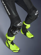 Kombinezon skórzany Alpinestars GP Force V2, jednoczęściowy czarny kombinezon skórzany, zbliżenie na nogi i kolana z białymi ślizgaczami z logo, harmonijkowe panele elastyczne, noszony z jaskrawymi neonowo-żółtymi butami wyścigowymi, wysokowydajny sprzęt do wyścigów motocyklowych
