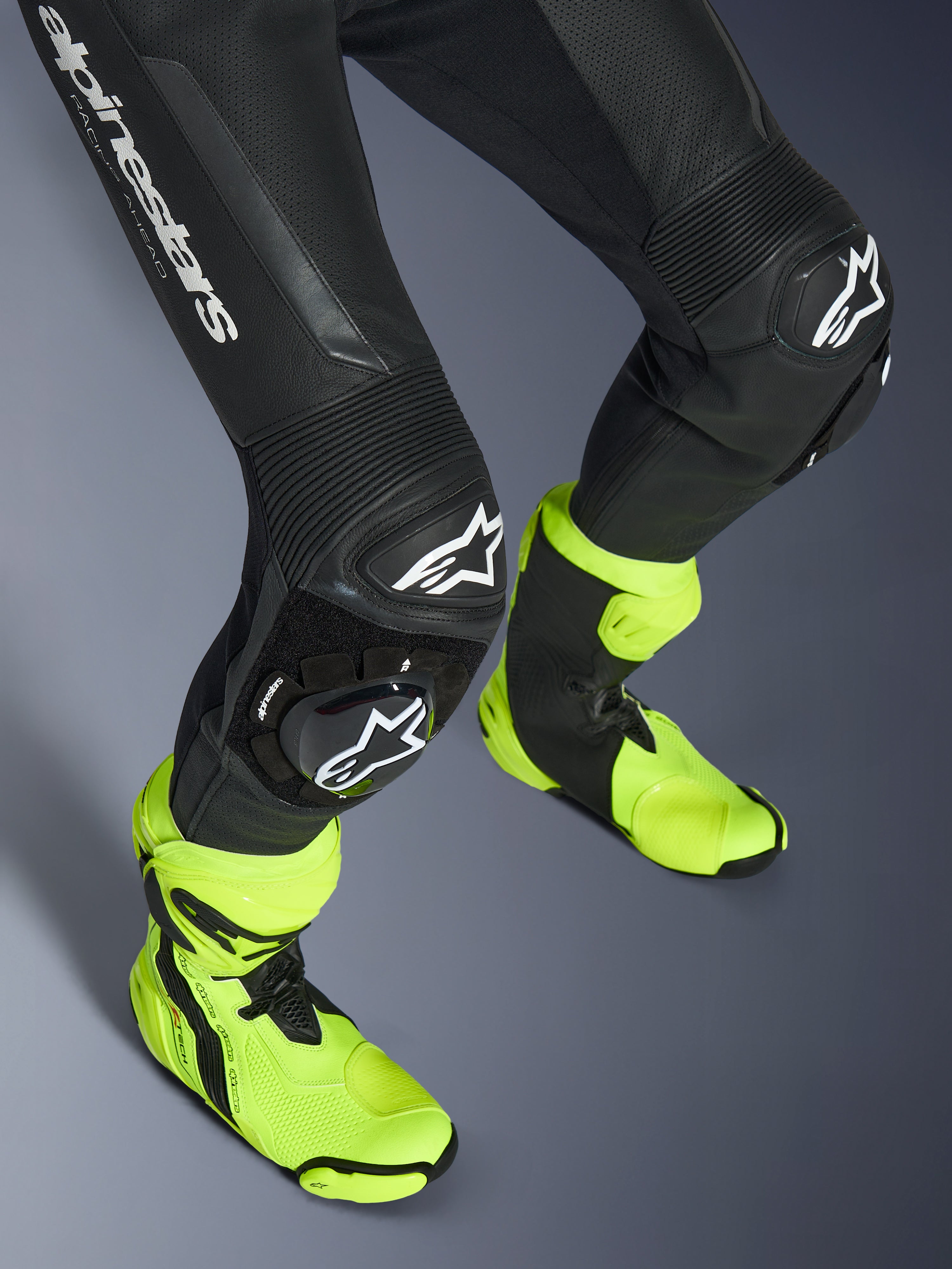 Kombinezon skórzany Alpinestars GP Force V2, jednoczęściowy czarny kombinezon skórzany, zbliżenie na nogi i kolana z białymi ślizgaczami z logo, harmonijkowe panele elastyczne, noszony z jaskrawymi neonowo-żółtymi butami wyścigowymi, wysokowydajny sprzęt do wyścigów motocyklowych