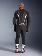Widok modelu od tyłu w jednoczęściowym kombinezonie skórzanym Alpinestars GP Force V2 w kolorze czarno-biało-czerwonym fluo, eksponujący aerodynamiczny garb i oznaczenie Tech-Air Ready, zestawiony z biało-czerwonymi profesjonalnymi butami wyścigowymi i czarnymi rękawicami.