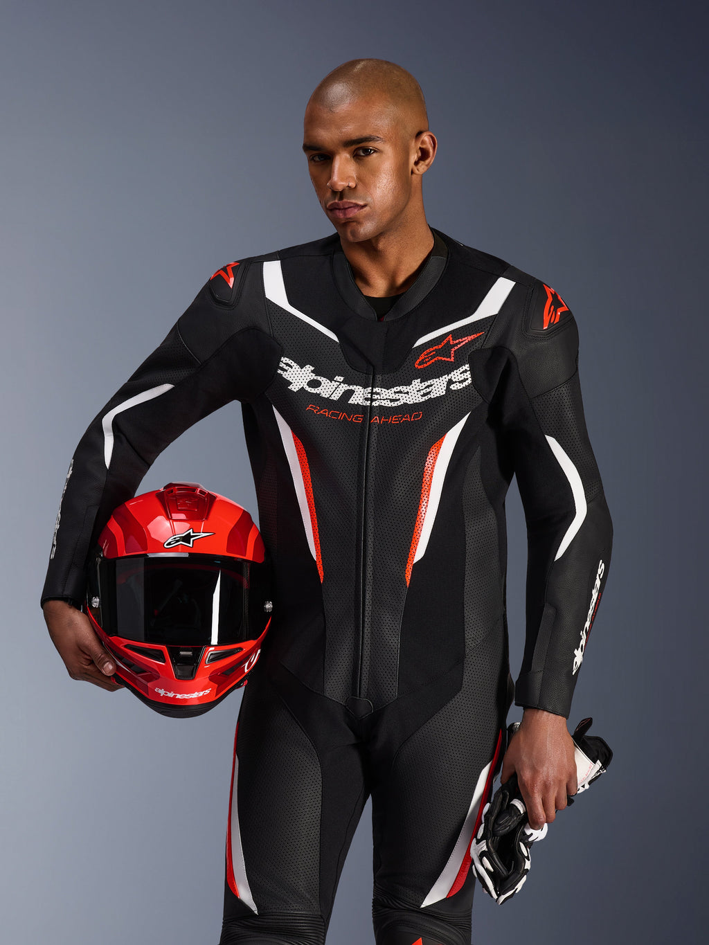 Alpinestars GP Force V2 Leather Suit, jednoczęściowy kombinezon motocyklowy, czarny, biały i czerwony fluo, prezentowany przez modela trzymającego czerwony kask i rękawice, sportowy design z perforowaną skórą i wyraźnym brandingiem