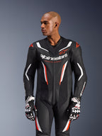 Jednoczęściowy kombinezon skórzany Alpinestars GP Force V2, motocyklowy kombinezon wyścigowy, czarno-biały z akcentami w kolorze czerwonym fluo, z perforowanymi panelami skórzanymi, wyraźnym logo na klatce piersiowej i napisem „Racing Ahead”, ergonomiczna konstrukcja ochronna, zestawiony z dopasowanymi rękawicami wyścigowymi.