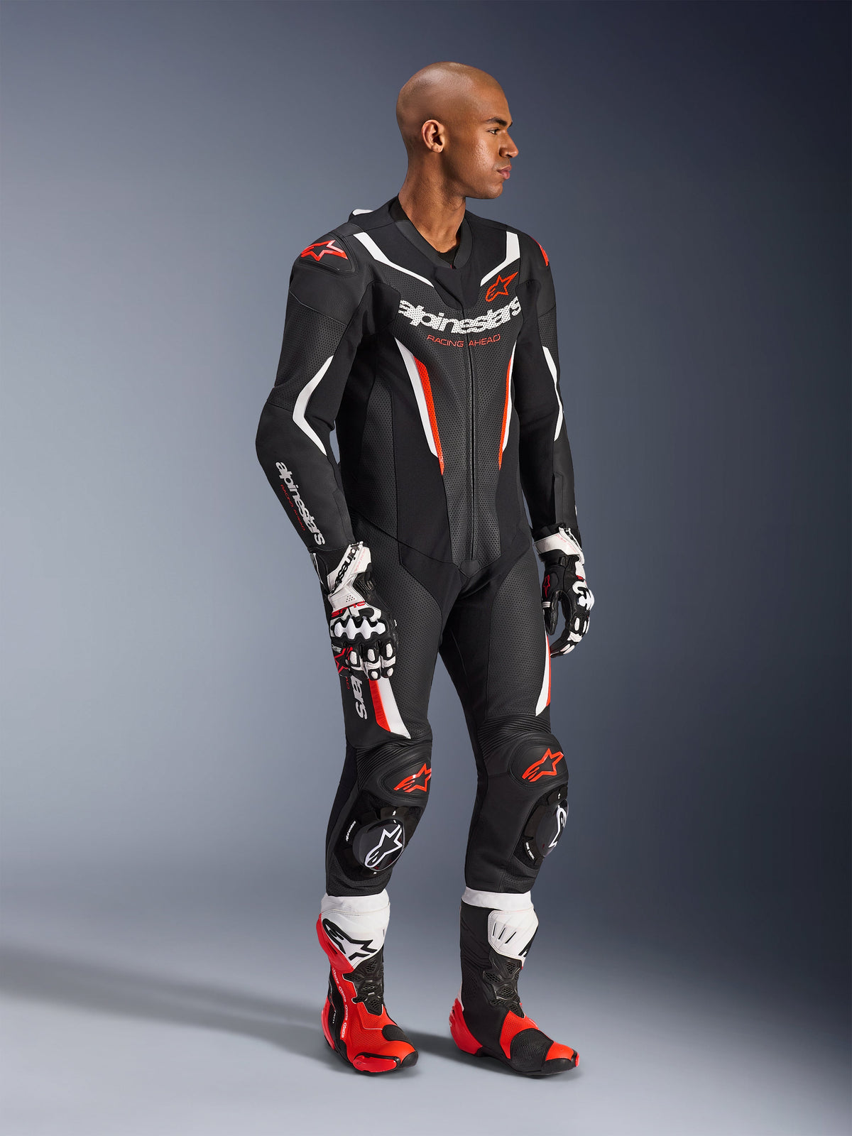 Model w jednoczęściowym skórzanym kombinezonie Alpinestars GP Force V2 w kolorze Black White Red Fluo, wysokowydajny sprzęt wyścigowy z perforowanymi panelami i osłonami DFS Lite, zestawiony z czerwono-białymi butami i rękawicami wyścigowymi, profesjonalna estetyka motorsportowa