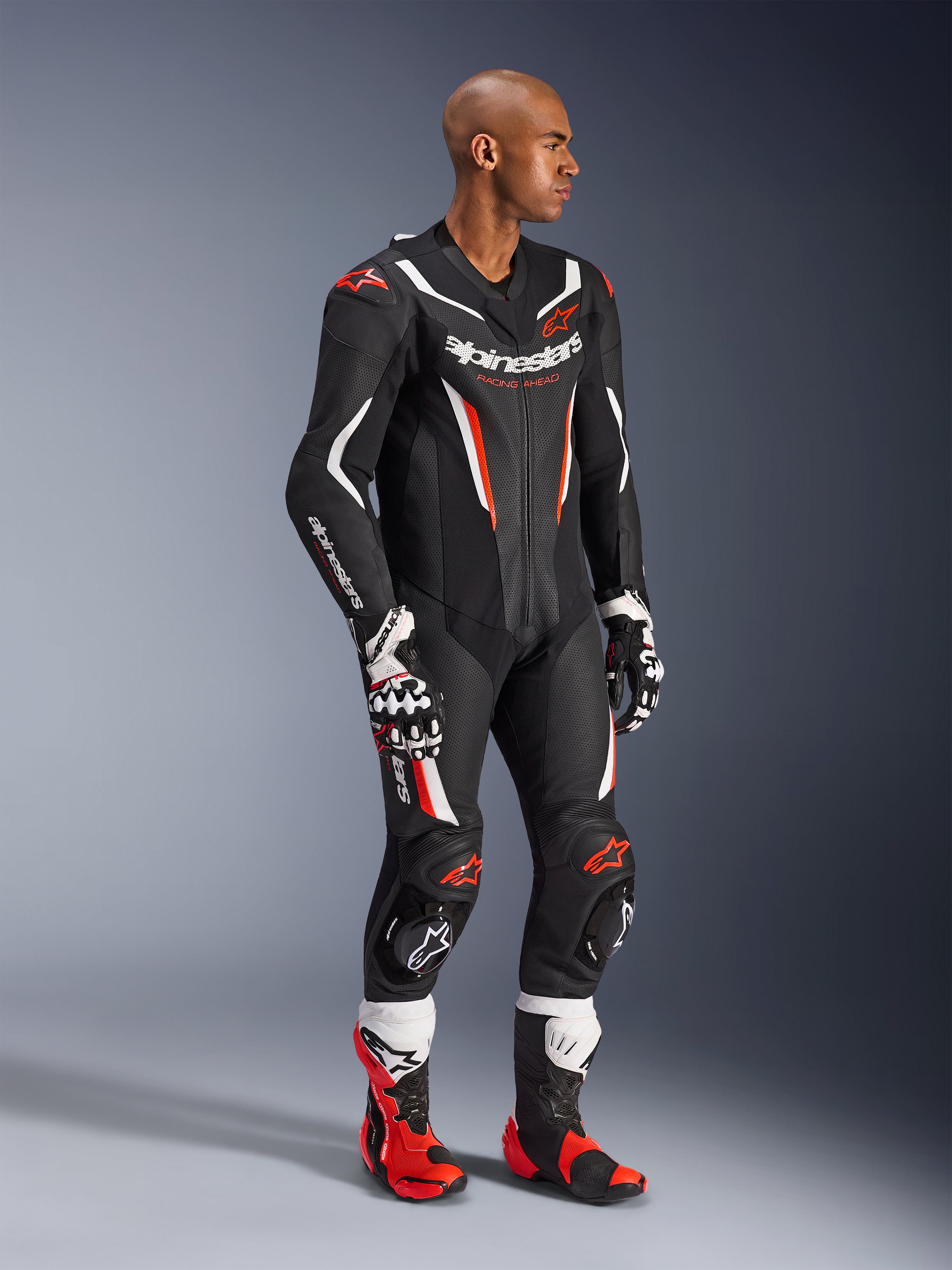 Model w jednoczęściowym skórzanym kombinezonie Alpinestars GP Force V2 w kolorze Black White Red Fluo, wysokowydajny sprzęt wyścigowy z perforowanymi panelami i osłonami DFS Lite, zestawiony z czerwono-białymi butami i rękawicami wyścigowymi, profesjonalna estetyka motorsportowa