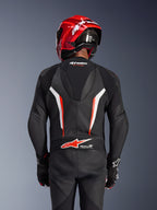 Alpinestars GP Force V2 Leather Suit 1-Piece, czarno-biało-czerwony fluo, widok od tyłu motocyklisty w czerwonym kasku wyścigowym i rękawicach, perforowane panele skórzane, aerodynamiczny garb z logo Alpinestars, logo Tech-Air Ready w dolnej części pleców, profesjonalny kombinezon wyścigowy.