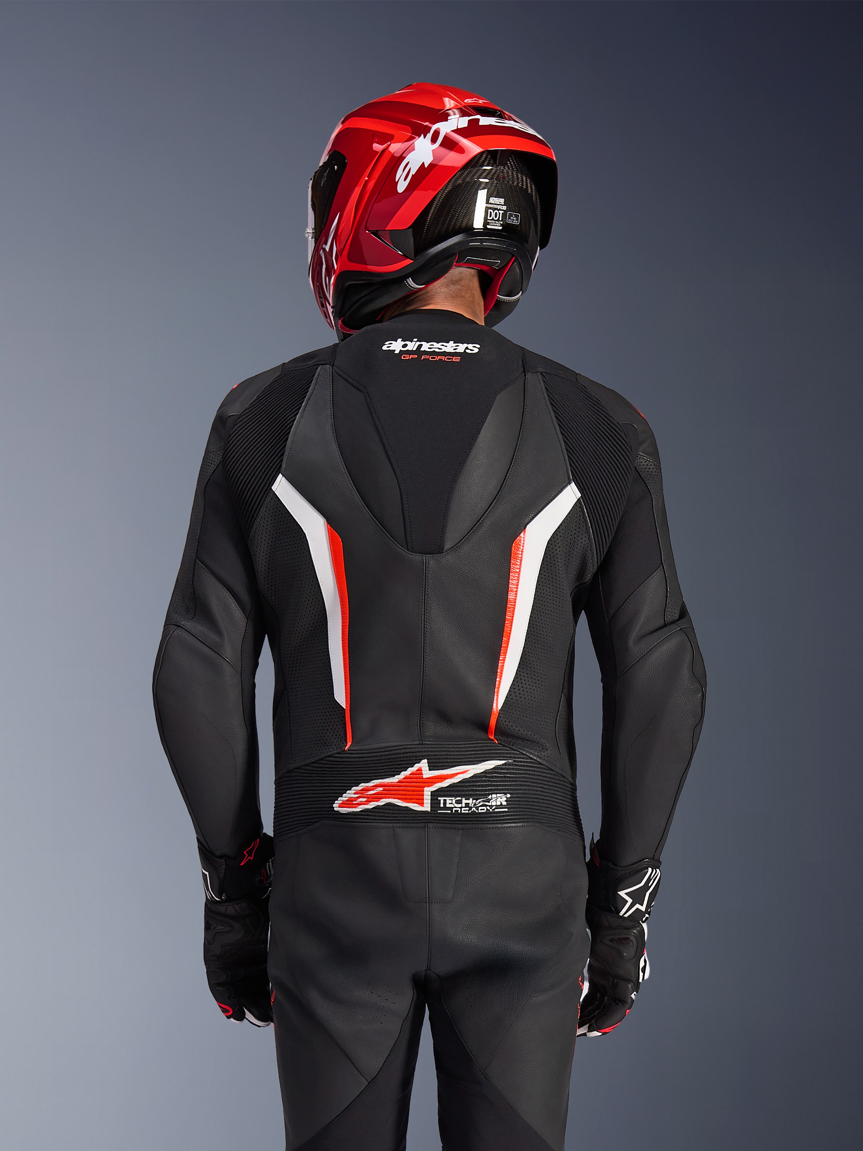 Alpinestars GP Force V2 Leather Suit 1-Piece, czarno-biało-czerwony fluo, widok od tyłu motocyklisty w czerwonym kasku wyścigowym i rękawicach, perforowane panele skórzane, aerodynamiczny garb z logo Alpinestars, logo Tech-Air Ready w dolnej części pleców, profesjonalny kombinezon wyścigowy.