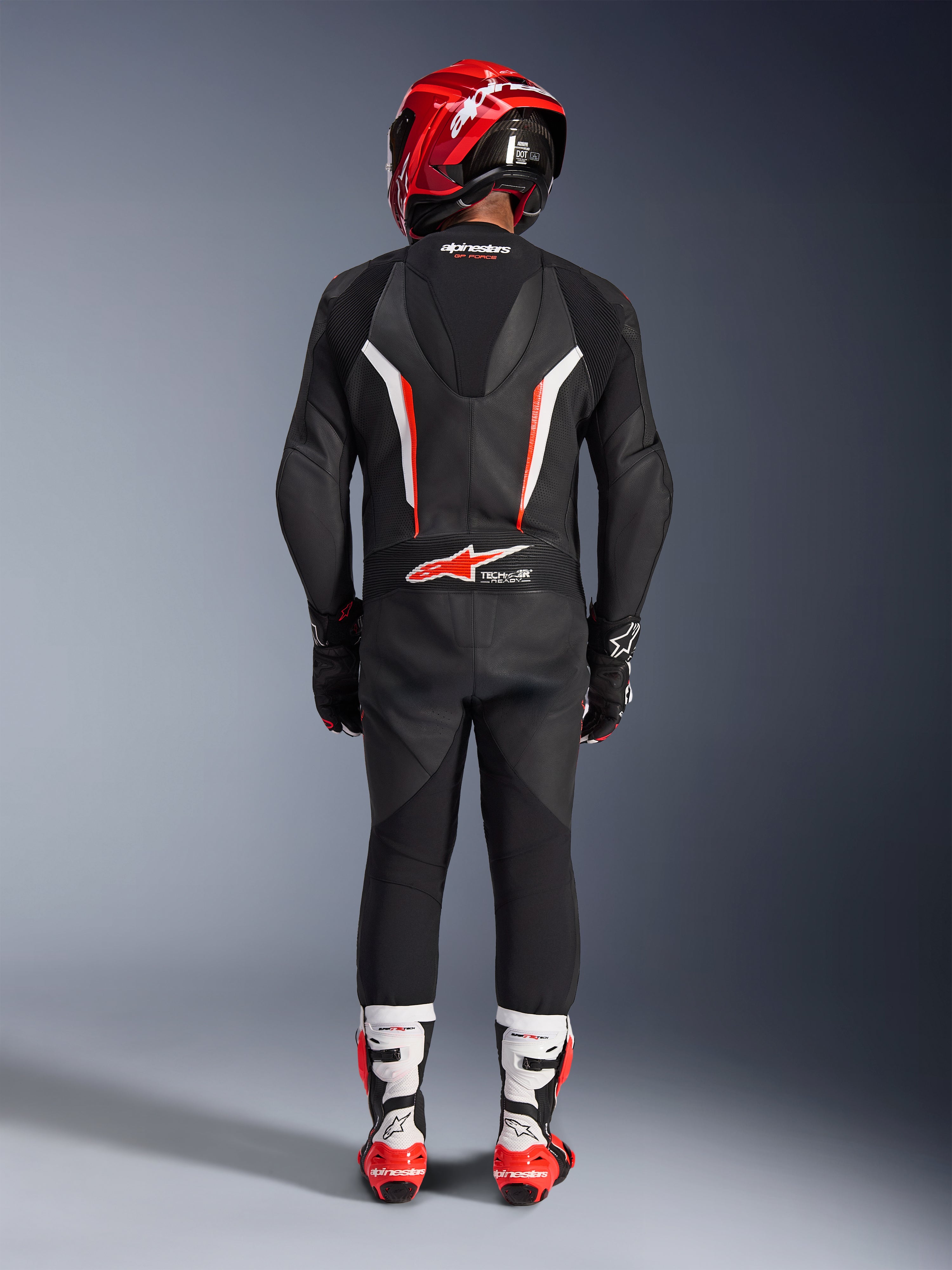 Motocyklista widziany od tyłu w jednoczęściowym skórzanym kombinezonie Alpinestars GP Force V2 w kolorystyce Black White Red Fluo, z aerodynamicznym garbem i oznaczeniem Tech-Air Ready, zestawiony z czerwonym kaskiem wyścigowym oraz biało-czerwonymi butami, profesjonalny design odzieży torowej.