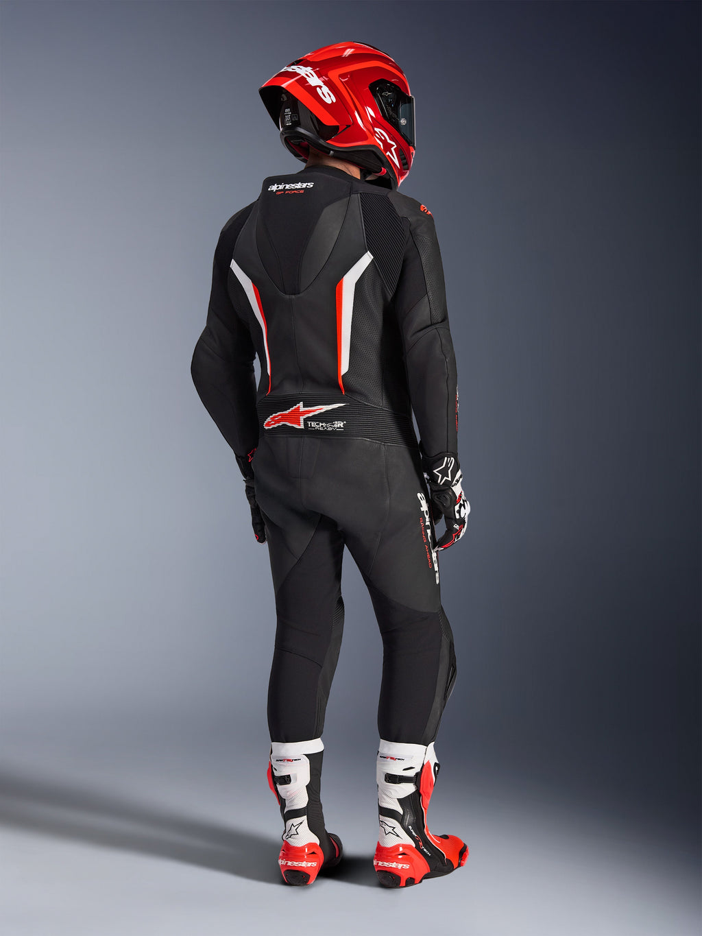Motocyklista widziany od tyłu w jednoczęściowym skórzanym kombinezonie Alpinestars GP Force V2 w kolorze czarnym, białym i czerwonym fluo, z aerodynamicznym garbem i perforowanymi panelami skórzanymi, w zestawie z czerwonym kaskiem Alpinestars, rękawicami wyścigowymi oraz czerwono-białymi butami motocyklowymi, profesjonalny ubiór wyścigowy.