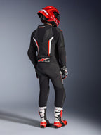 Motocyklista widziany od tyłu w jednoczęściowym skórzanym kombinezonie Alpinestars GP Force V2 w kolorze czarnym, białym i czerwonym fluo, z aerodynamicznym garbem i perforowanymi panelami skórzanymi, w zestawie z czerwonym kaskiem Alpinestars, rękawicami wyścigowymi oraz czerwono-białymi butami motocyklowymi, profesjonalny ubiór wyścigowy.