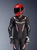 Motocyklista w jednoczęściowym kombinezonie skórzanym Alpinestars GP Force V2 w kolorystyce czarno-biało-czerwonej fluo, z dopasowanym czerwonym kaskiem wyścigowym i rękawicami, posiada perforowane panele skórzane i wyraźny branding, profesjonalny sprzęt wyścigowy