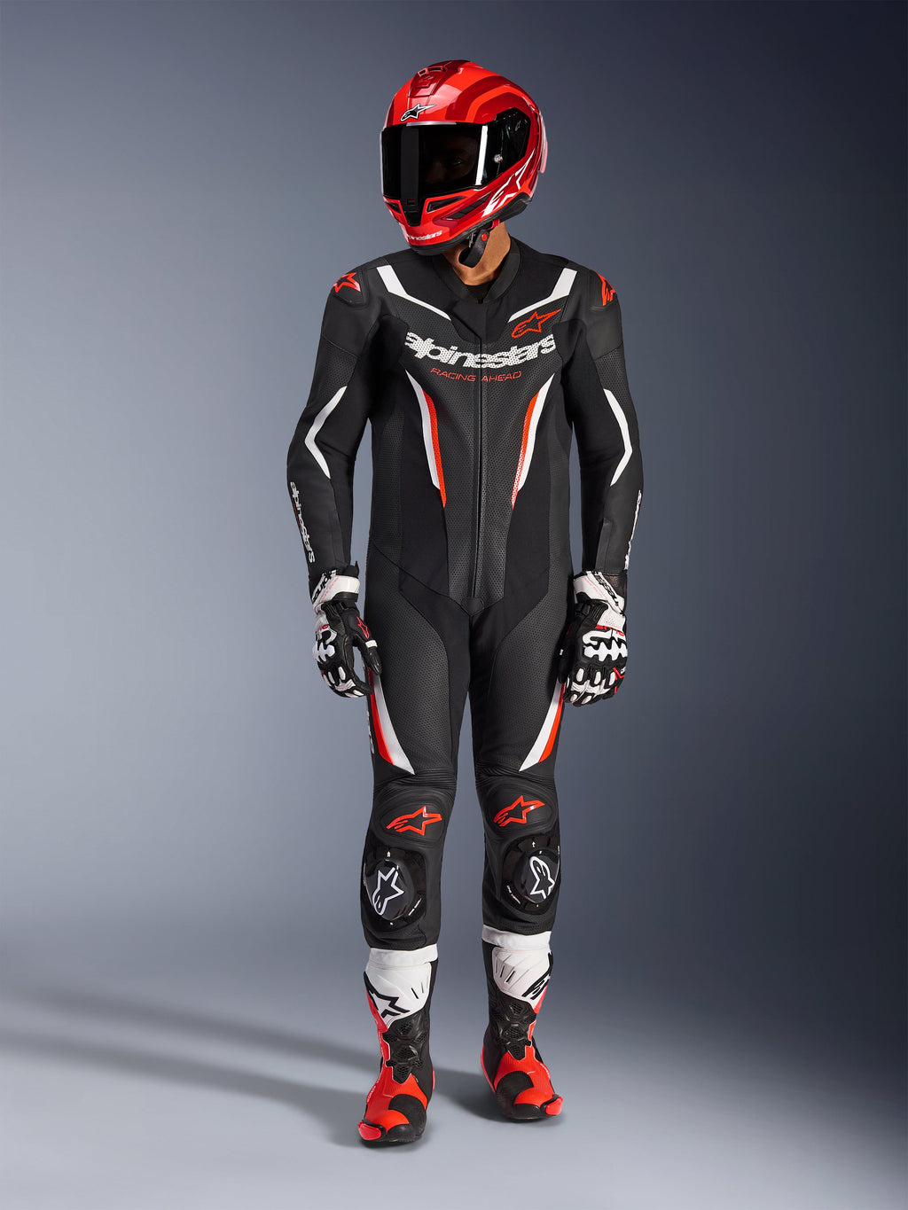 Kombinezon skórzany Alpinestars GP Force V2 - 1-częściowy, czarny, biały i czerwony fluo, zestawiony z czerwonym kaskiem wyścigowym, czarno-białymi rękawicami i czerwono-czarnymi butami, posiada perforowane panele skórzane i branding Alpinestars, profesjonalna odzież motocyklowa wyścigowa