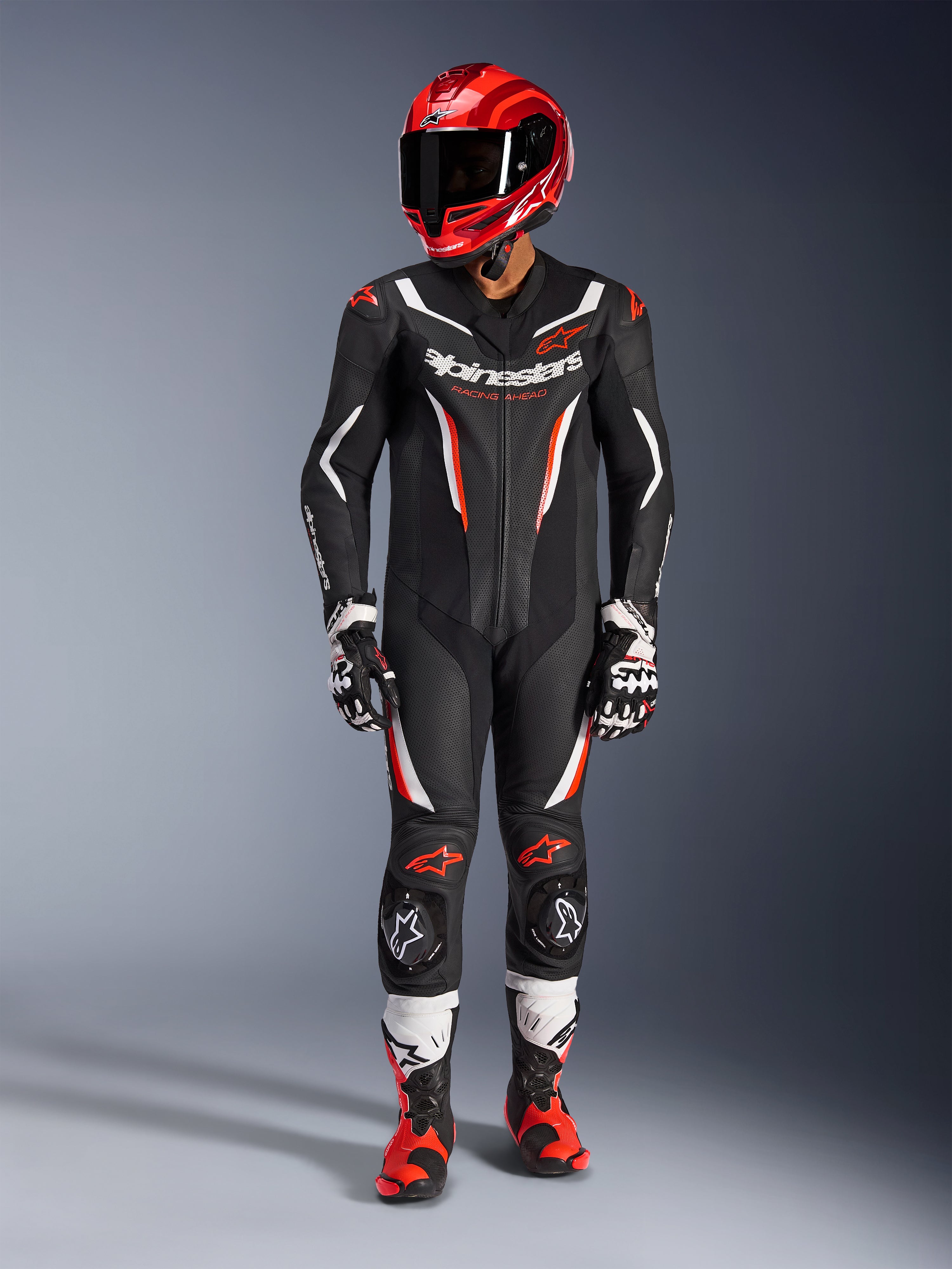 Kombinezon skórzany Alpinestars GP Force V2 - 1-częściowy, czarny, biały i czerwony fluo, zestawiony z czerwonym kaskiem wyścigowym, czarno-białymi rękawicami i czerwono-czarnymi butami, posiada perforowane panele skórzane i branding Alpinestars, profesjonalna odzież motocyklowa wyścigowa