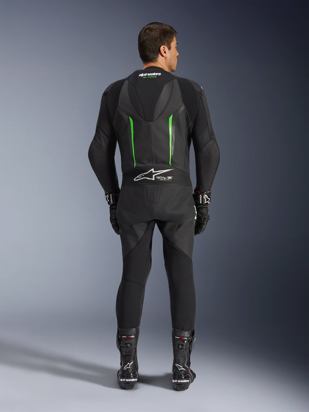 Widok z tyłu jednoczęściowego kombinezonu skórzanego Alpinestars GP Force V2 w kolorze czarno-zielonym, z aerodynamicznym garbem, perforowanymi panelami i logo Tech-Air Ready w dolnej części pleców, noszony z czarnymi rękawicami i butami wyścigowymi.