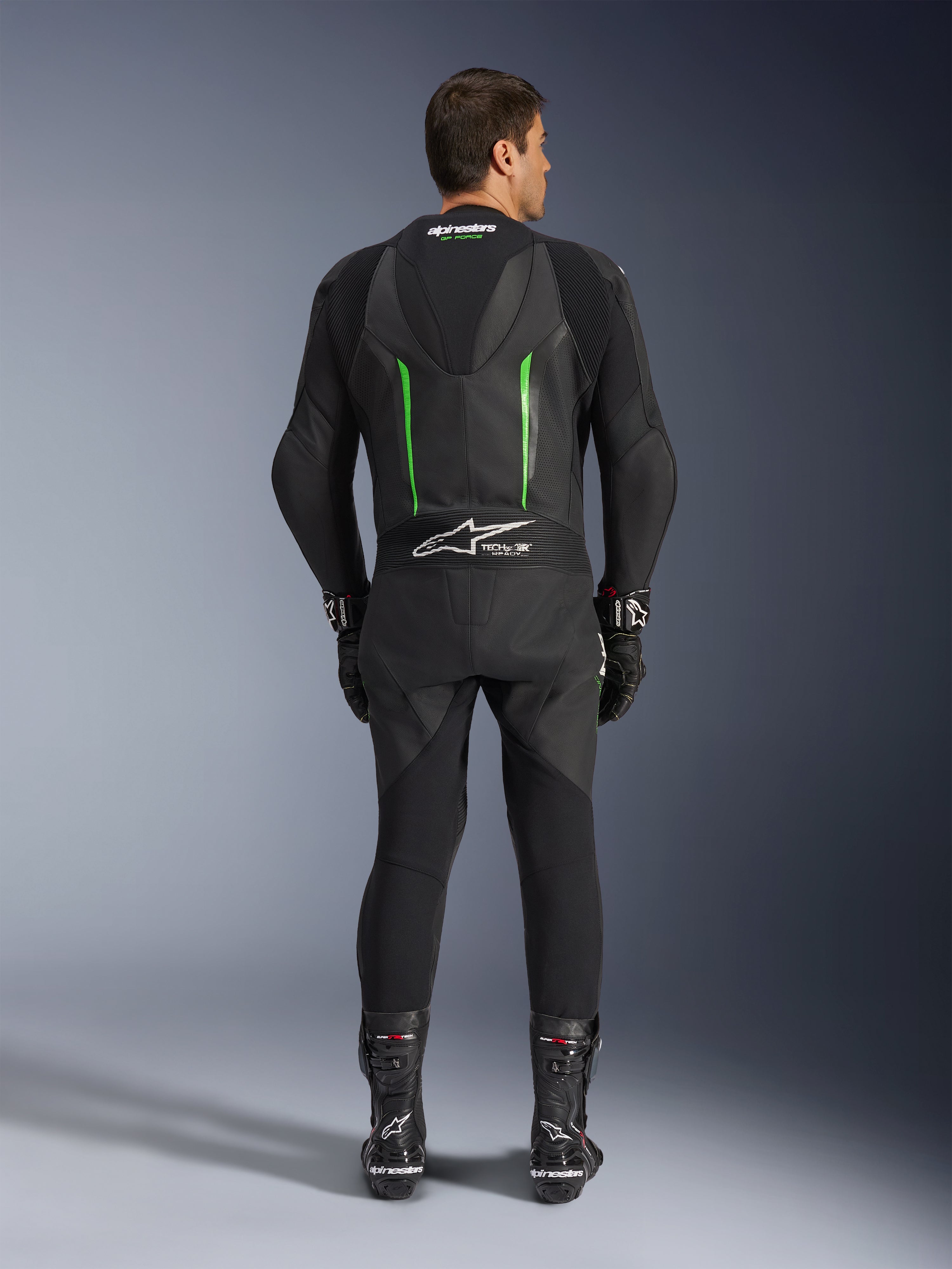 Widok z tyłu jednoczęściowego kombinezonu skórzanego Alpinestars GP Force V2 w kolorze czarno-zielonym, z aerodynamicznym garbem, perforowanymi panelami i logo Tech-Air Ready w dolnej części pleców, noszony z czarnymi rękawicami i butami wyścigowymi.