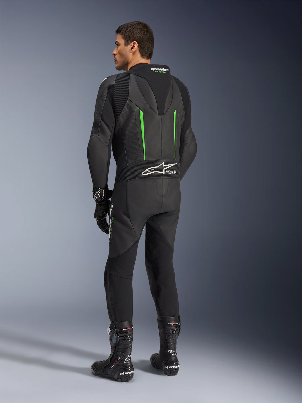 Skórzany kombinezon Alpinestars GP Force V2, jednoczęściowy kombinezon motocyklowy, czarny z zielonymi akcentami, widok z tyłu pokazujący aerodynamiczny garb i białe logo na harmonijkowym panelu elastycznym, w zestawieniu z czarnymi butami i rękawicami wyścigowymi.