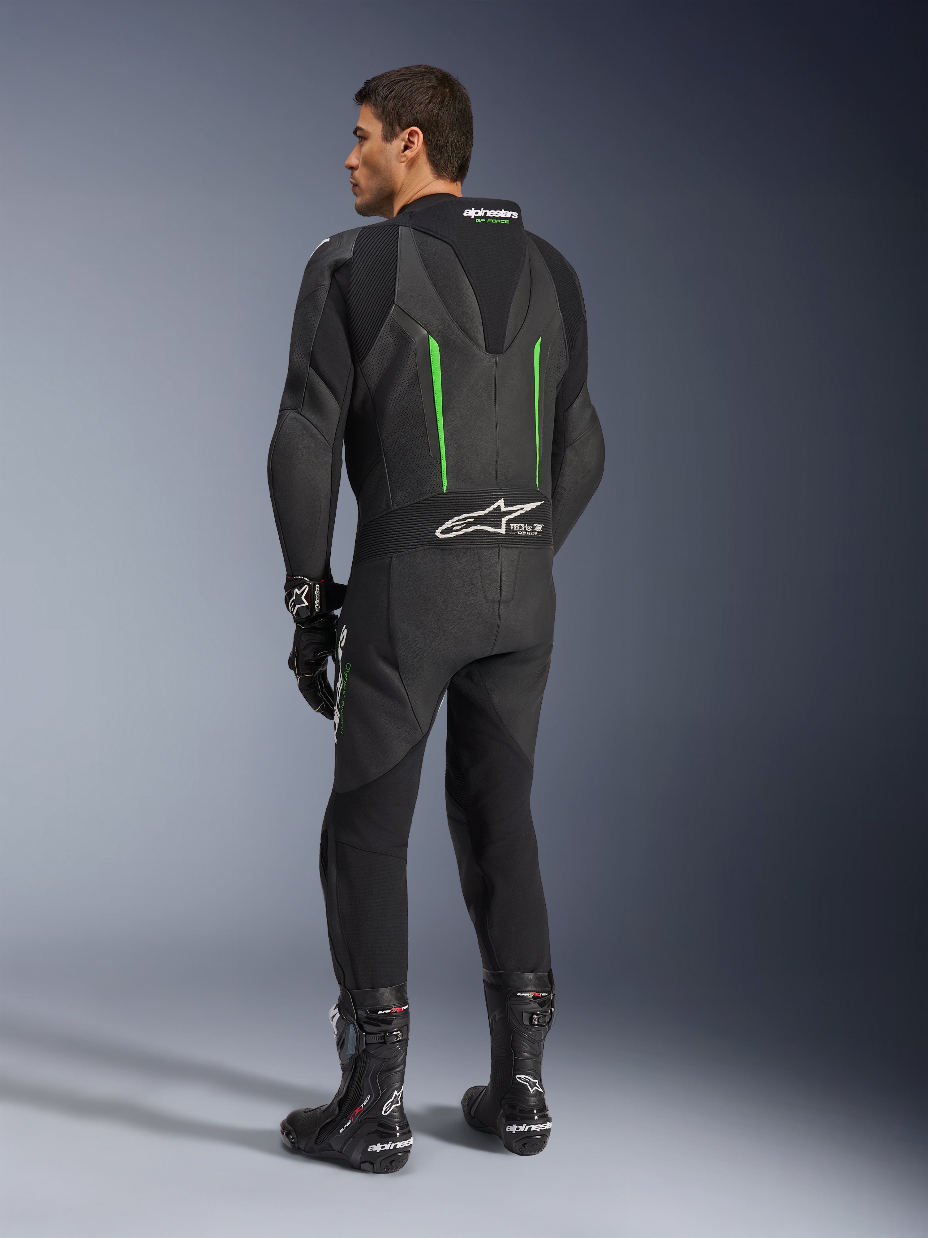 Skórzany kombinezon Alpinestars GP Force V2, jednoczęściowy kombinezon motocyklowy, czarny z zielonymi akcentami, widok z tyłu pokazujący aerodynamiczny garb i białe logo na harmonijkowym panelu elastycznym, w zestawieniu z czarnymi butami i rękawicami wyścigowymi.