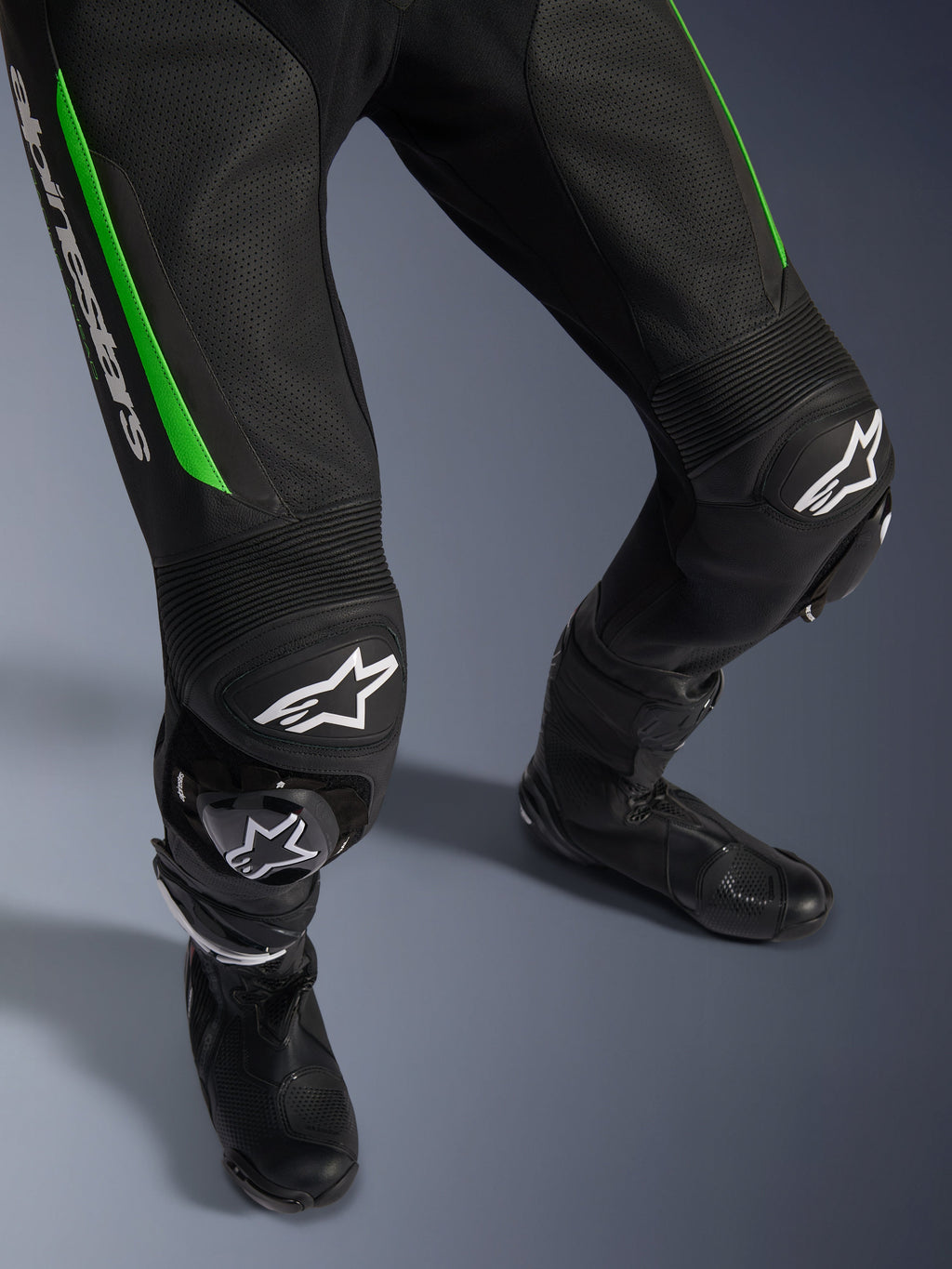 Alpinestars GP Force V2 Leather Suit - 1Piece, zbliżenie na nogi w kolorze czarno-zielonym, perforowana skórzana konstrukcja, harmonijkowe panele elastyczne na kolanach, białe logo Alpinestars i zintegrowane slidery kolan, noszony z czarnymi butami wyścigowymi