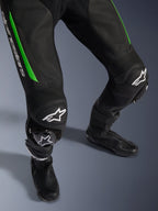 Alpinestars GP Force V2 Leather Suit - 1Piece, zbliżenie na nogi w kolorze czarno-zielonym, perforowana skórzana konstrukcja, harmonijkowe panele elastyczne na kolanach, białe logo Alpinestars i zintegrowane slidery kolan, noszony z czarnymi butami wyścigowymi