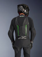 Widok z tyłu motocyklisty w skórzanym kombinezonie Alpinestars GP Force V2 w kolorze Black Green, z aerodynamicznym garbem, oznaczeniem Tech-Air Ready na harmonijkowym panelu w dolnej części pleców i konstrukcją z perforowanej skóry, w czarnym wyścigowym kasku z włókna węglowego.